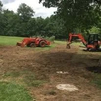 Construction site with a red tractor and a mini excavator on dirt, surrounded by green trees.