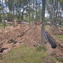 Construction site with trees and a large dug trench, possibly for utility work or drainage, with construction vehicles in the background.