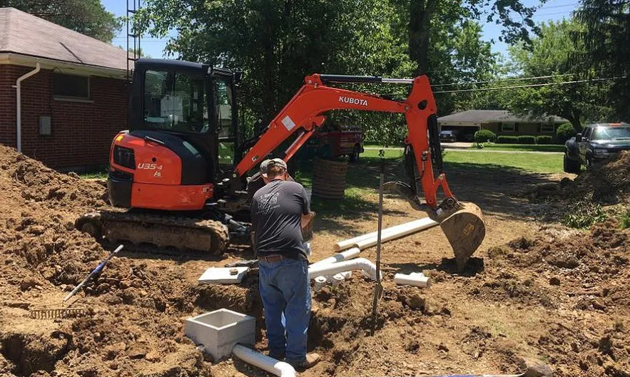 Man working on underground pipe installation using a small orange Kubota excavator in a residential yard.