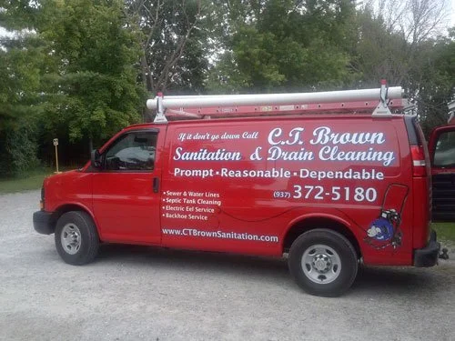 Red sanitation and drain cleaning service van with the company name C.T. Brown Sanitation & Drain Cleaning, phone number 937-372-5180, and services listed including sewer and water lines, septic tank cleaning, electric eel service, and backhoe service, parked outdoors near green trees.