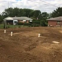 Empty plot of land with white markers, residential houses in the background, and a cloudy sky.