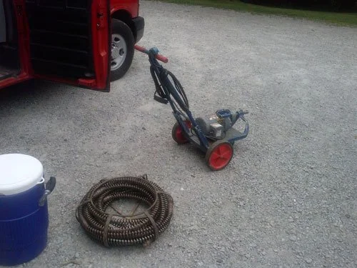 A lawn edging weed trimmer with a long handle and red wheels, a coiled black garden hose, and a blue cooler with a white lid on a gravel surface.