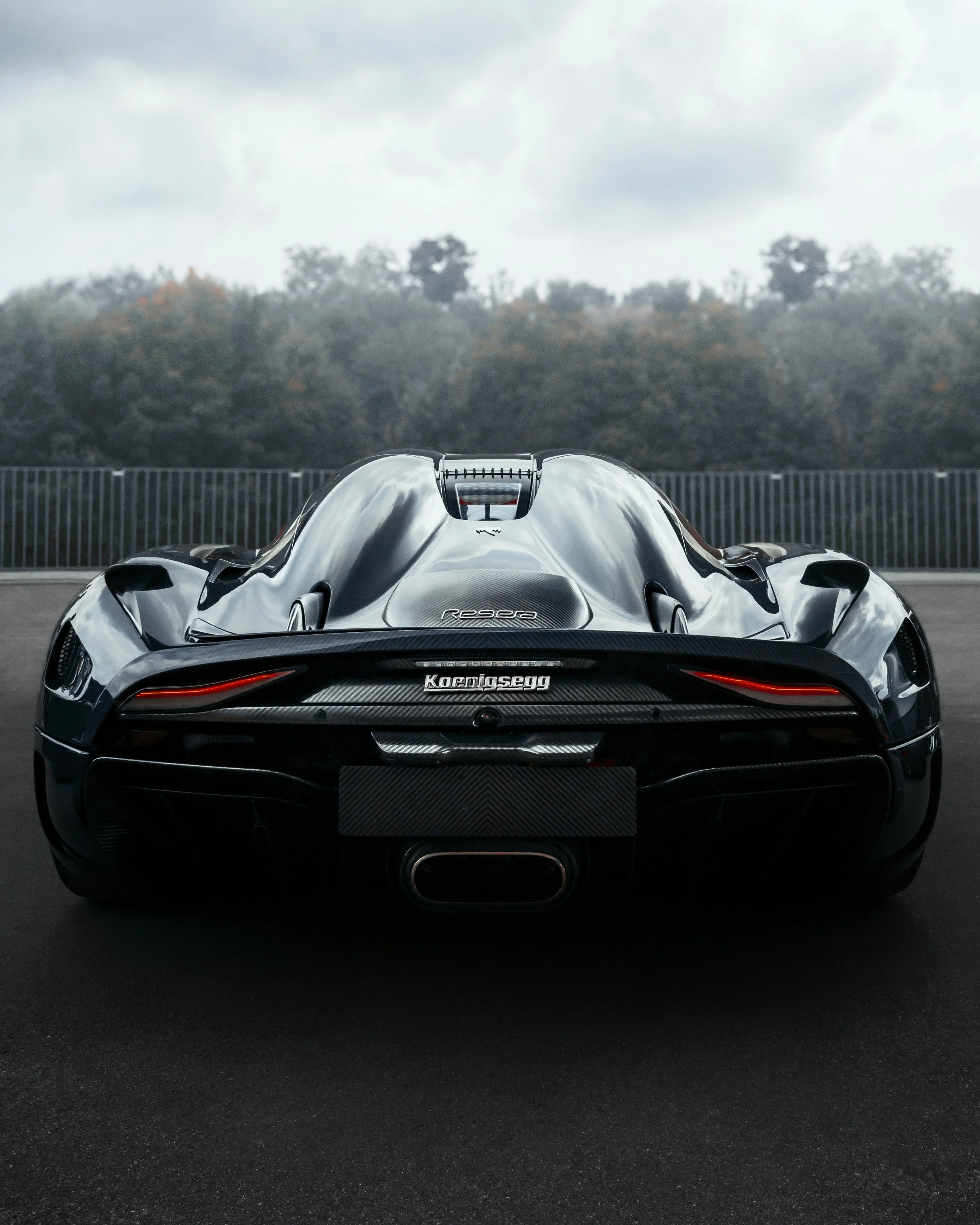 Rear view of a sleek, black Koenigsegg sports car parked on a dark asphalt surface, with a blurred natural background of trees and a cloudy sky.
