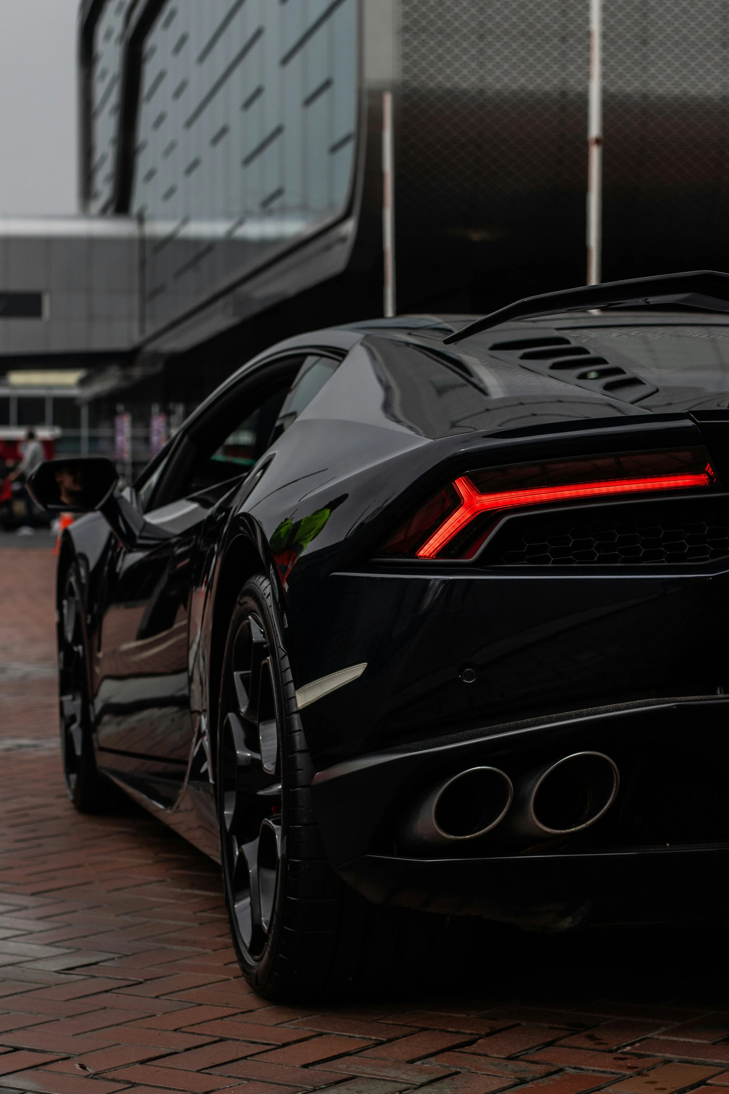 Close-up of the rear end of a black sports car with red tail lights, showing dual exhaust pipes, parked on a brick-paved surface. The background includes blurred buildings and a metal fence.