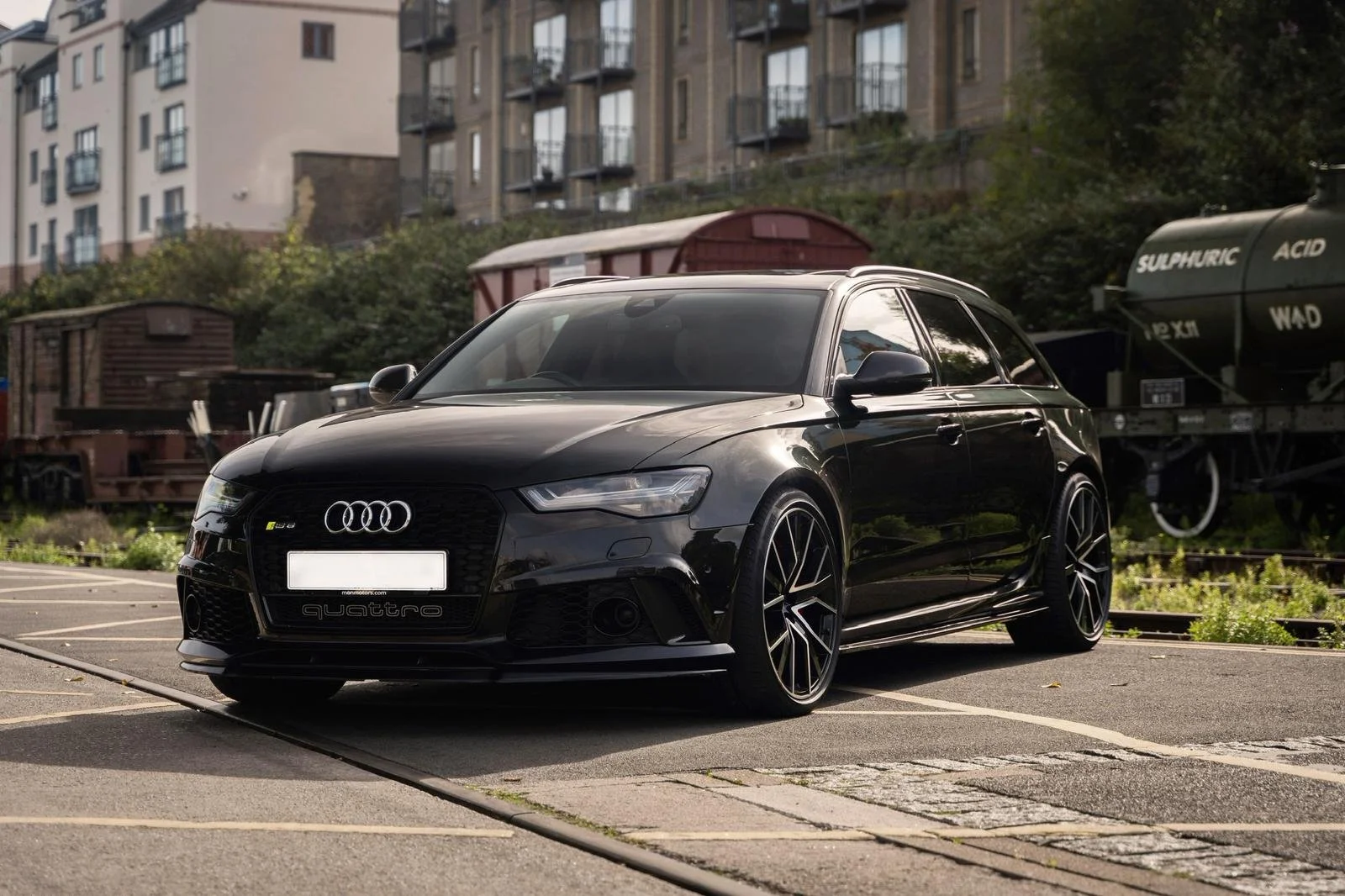 A sleek black Audi S6 parked in a parking lot, with a background of residential buildings and an old train car.