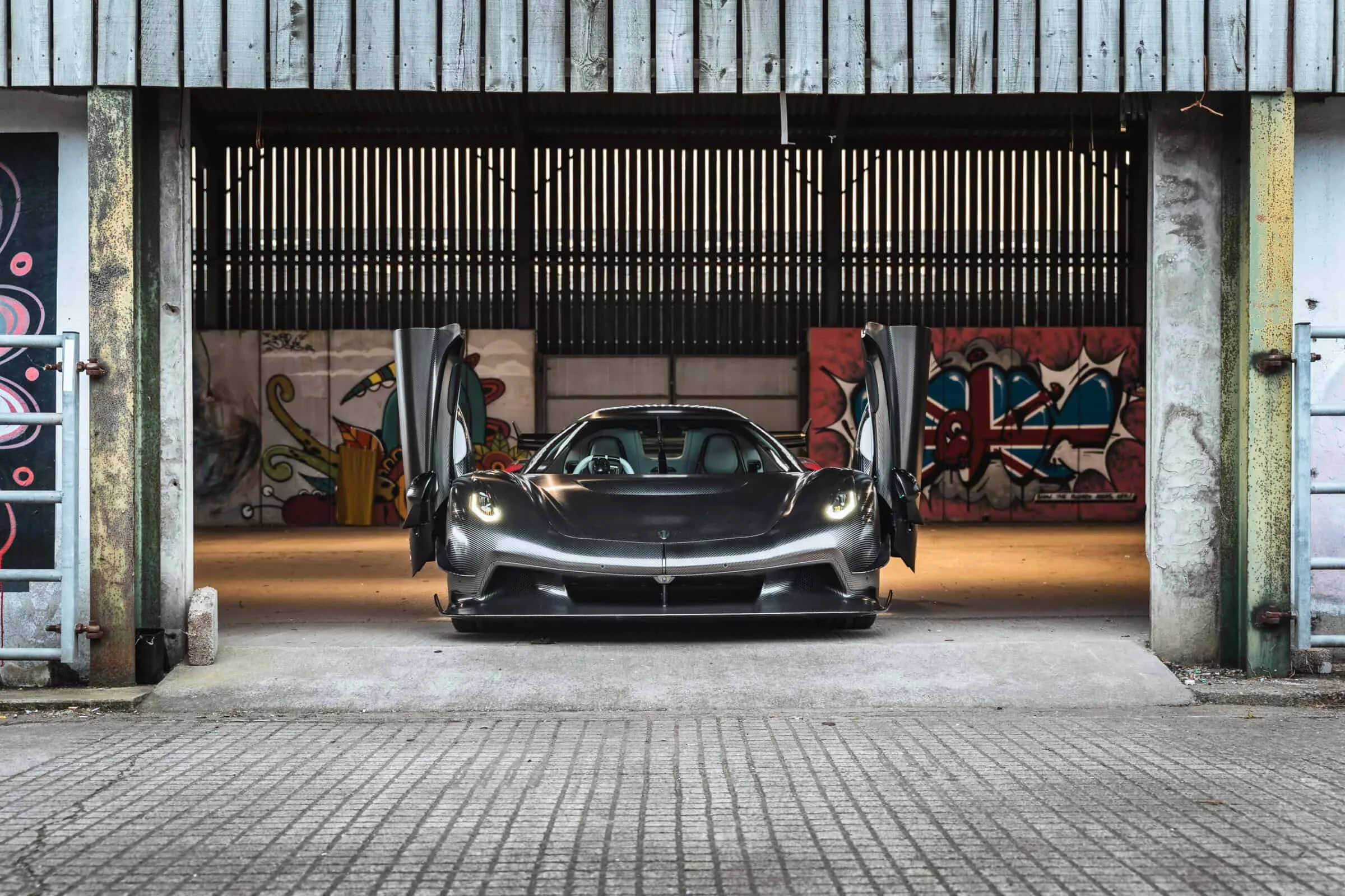 A sleek black sports car with gullwing doors open, parked inside an urban garage with graffiti art on the walls.