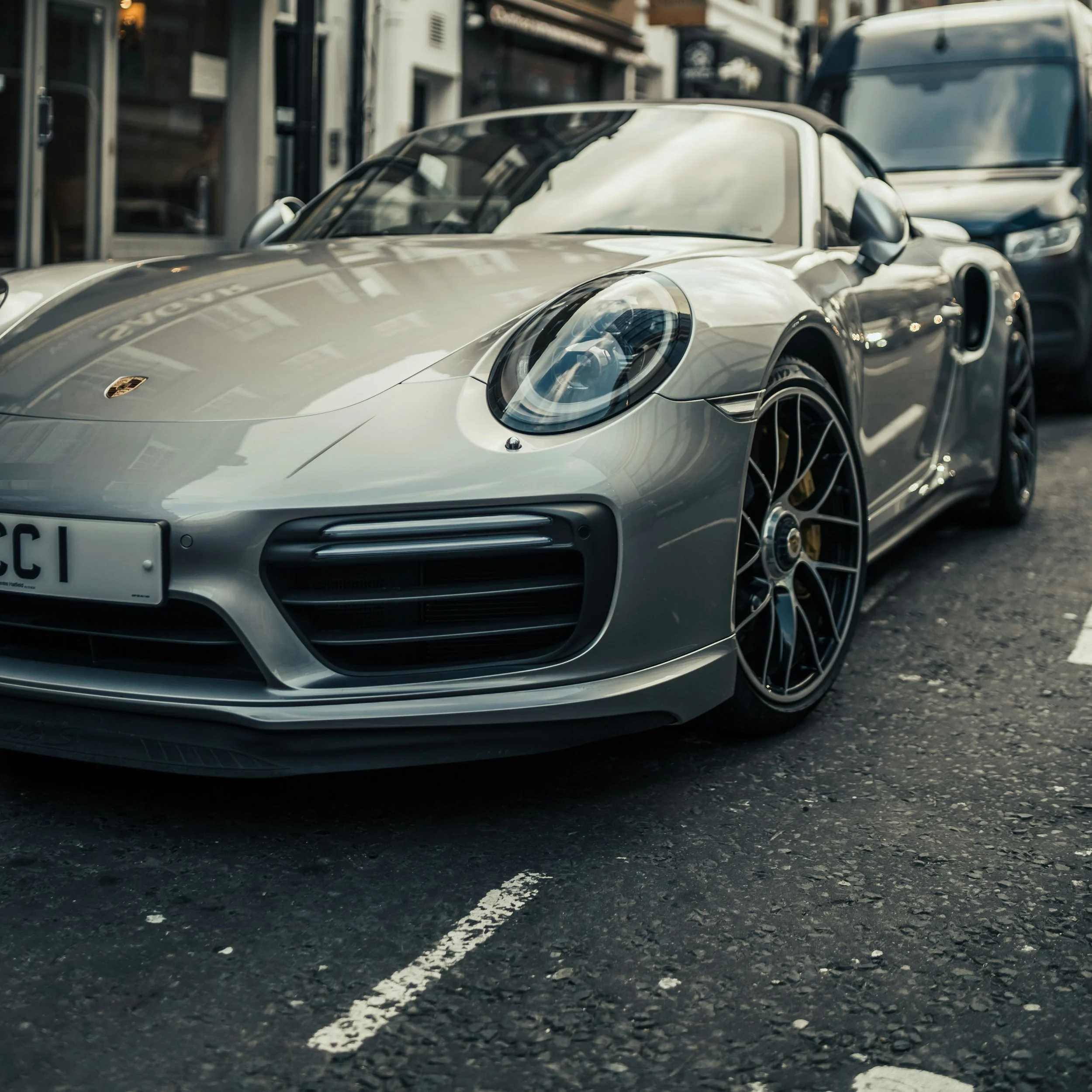 Close-up view of a silver Porsche sports car parked on a city street, with white dashed lines painted on the dark asphalt ground.