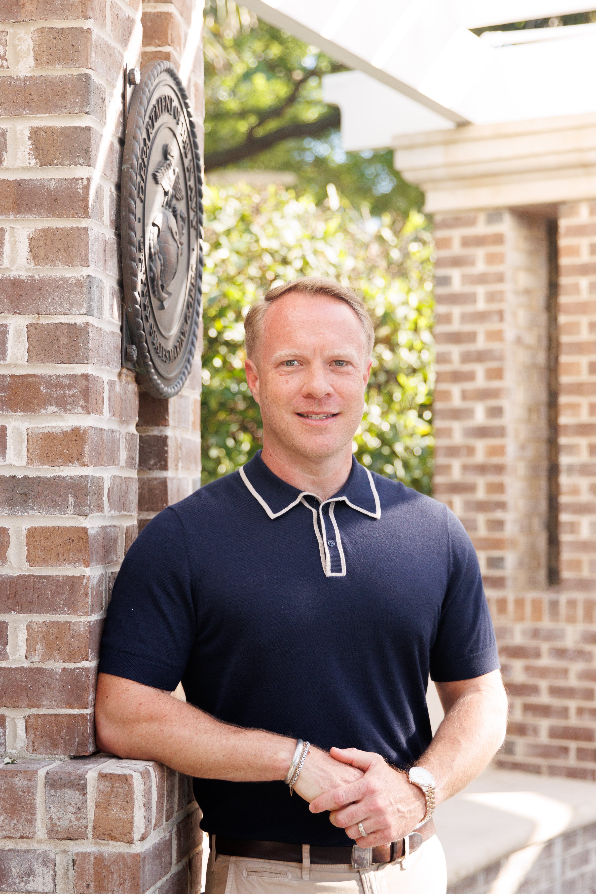 A man with short, light brown hair and wearing a navy polo shirt and beige pants standing outside next to a brick column. There is a metal emblem on the brick wall behind him, and greenery can be seen in the background.