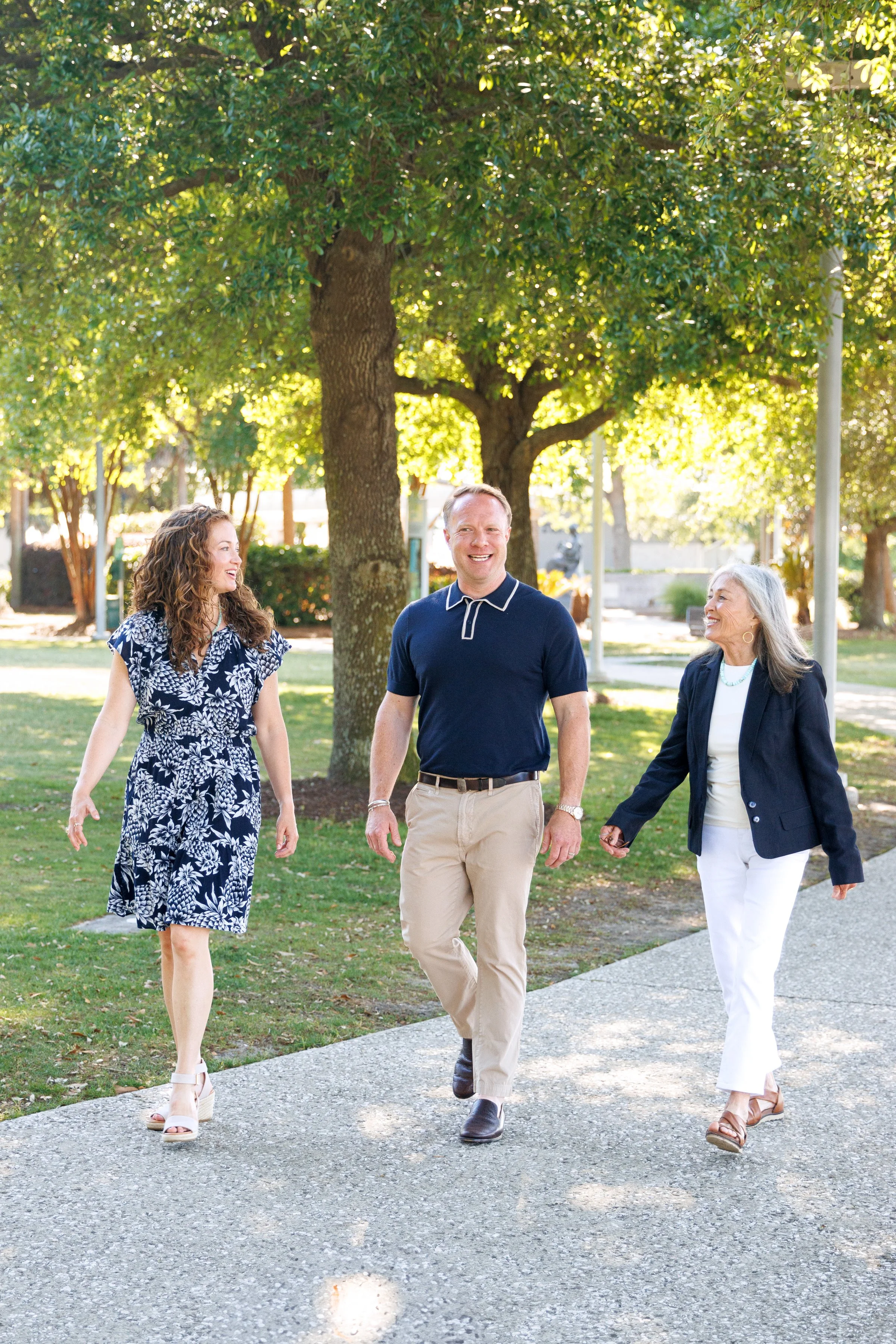 Three people, two women and one man, walking on a sidewalk in a park with green trees and sunlight filtering through the leaves, all smiling and engaged in conversation.