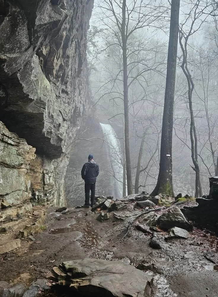 Person standing beside a misty forest waterfall beneath a rocky cliff