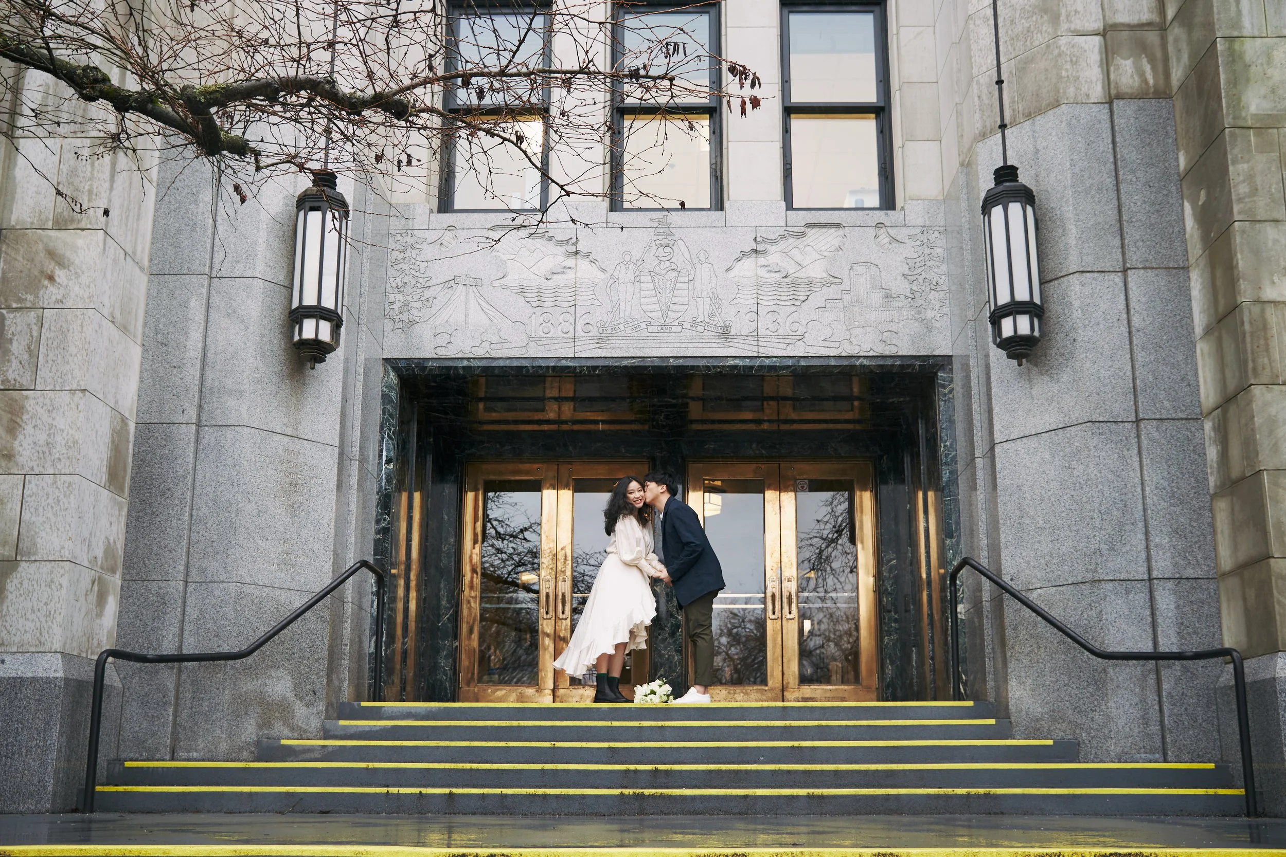 A bride and groom stand on the steps outside a stone building with large glass doors, holding hands and smiling at each other. The bride wears a white wedding dress and black boots, holding a bouquet of flowers, while the groom wears a dark suit. A b
