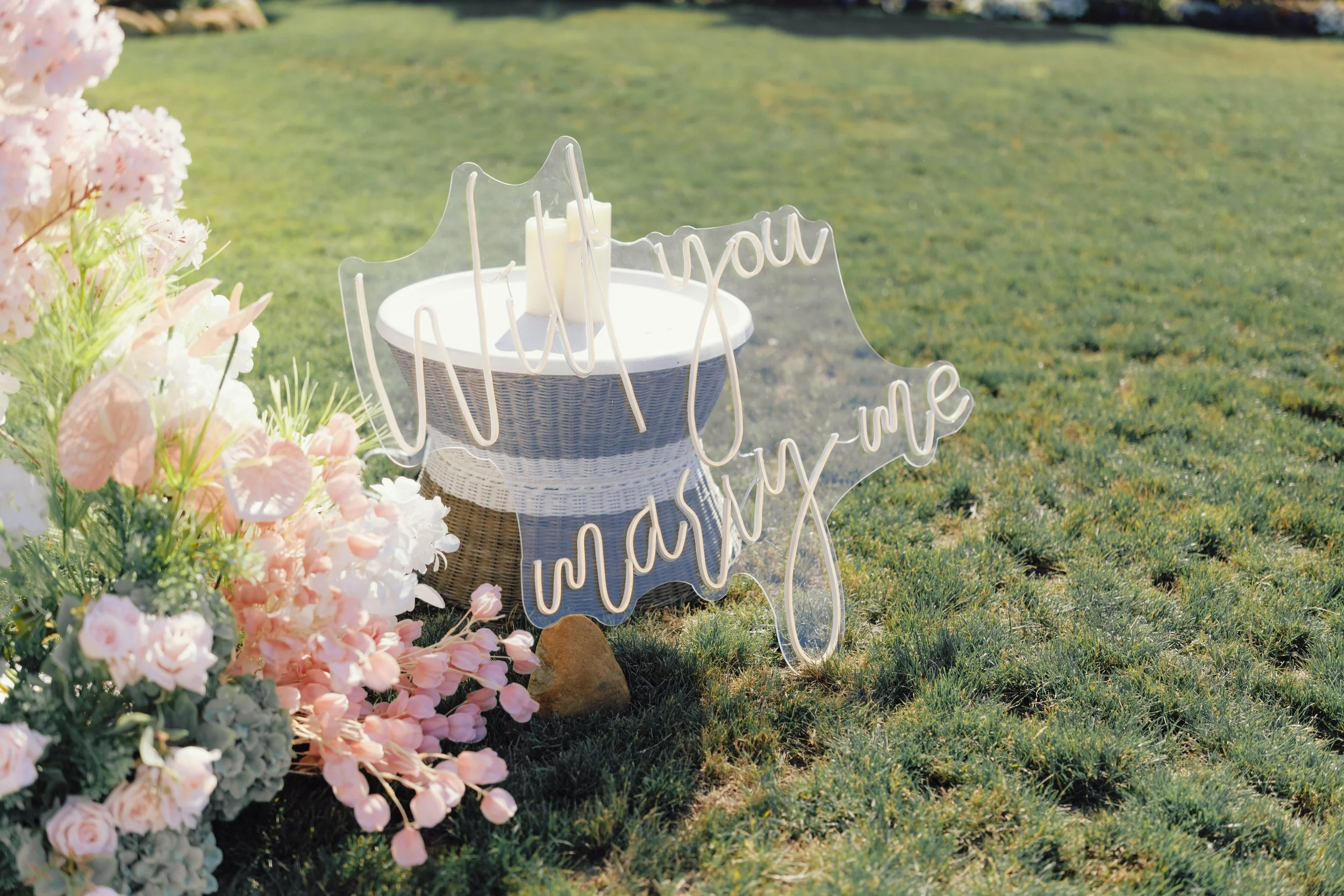 A wicker basket with a white cloth liner on lush green grass. There are pink and white flowers on the left side of the image. A clear acrylic sign with the words 'thank you' in cursive hangs on the basket.