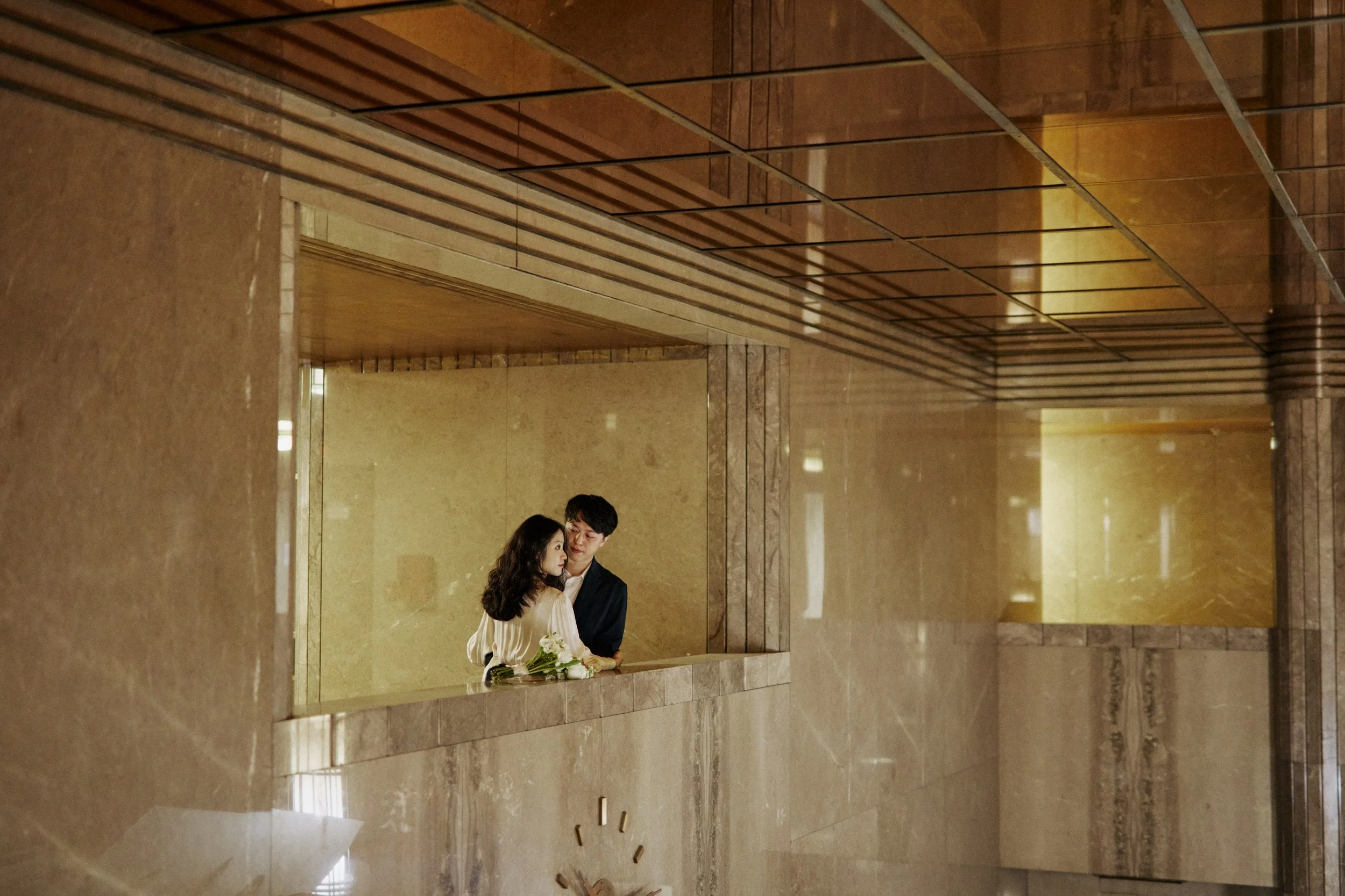 A couple dressed in formal attire, holding a bouquet of white flowers, standing in an elegant, marble-paneled room with warm lighting and reflective surfaces.