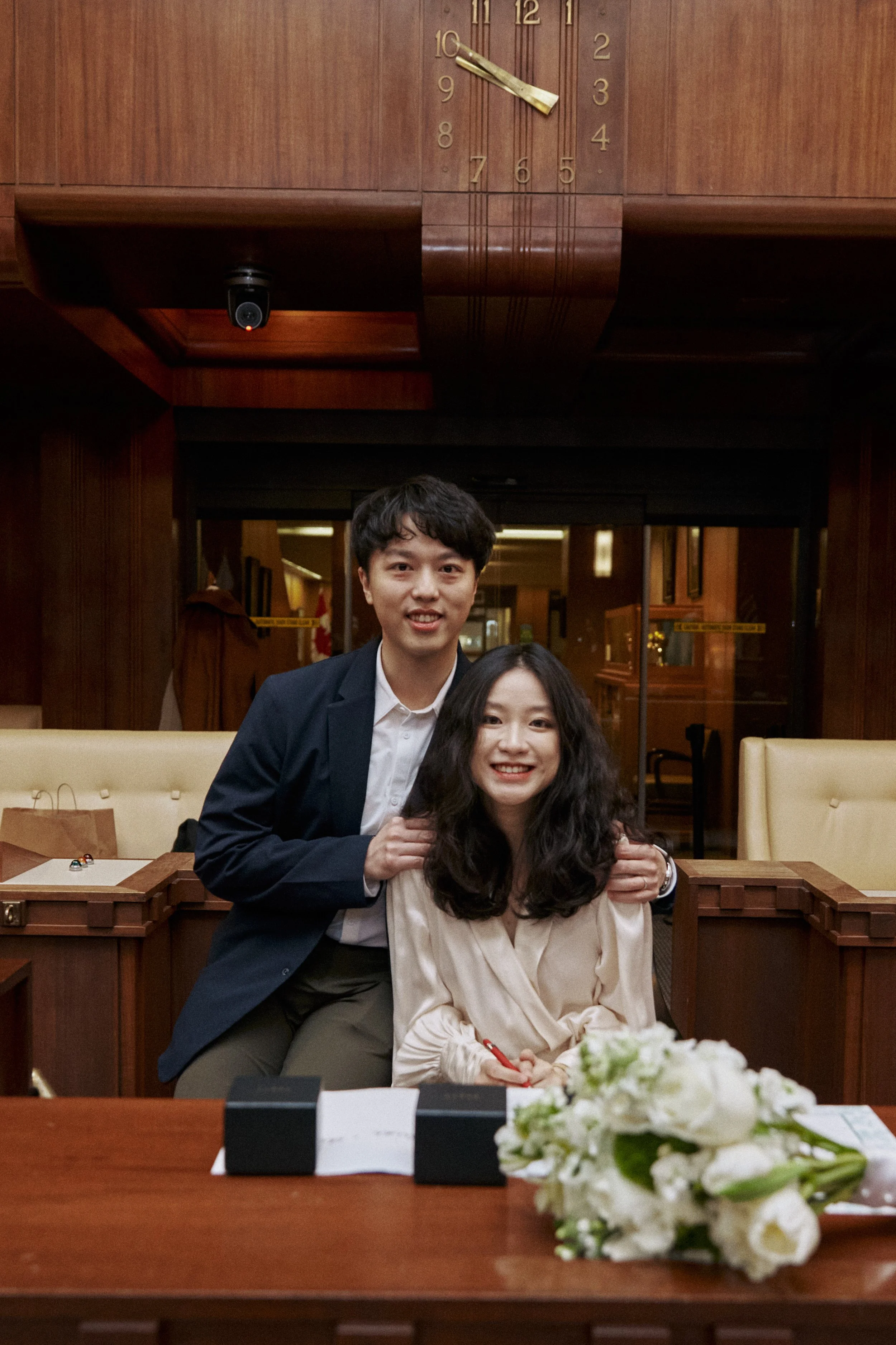 A man and a woman sitting at a table in a restaurant or banquet hall, smiling and posing for a photo. The table has a bouquet of white flowers and some documents or items placed on it. The background is wooden paneling with a clock on the wall showin