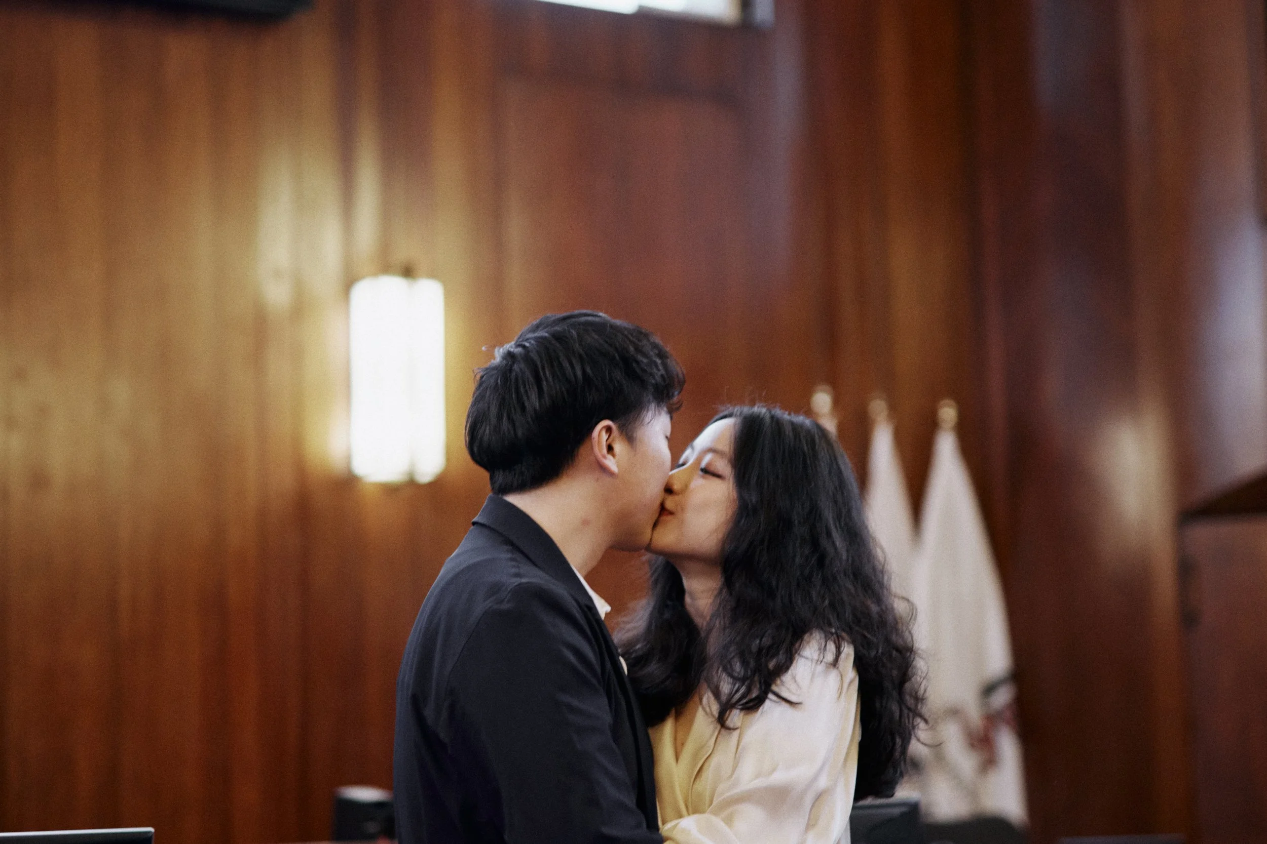 A young man and woman are sharing a kiss in a formal setting, possibly during a wedding or special ceremony, with the background featuring polished wooden paneling and white drapes or flags.