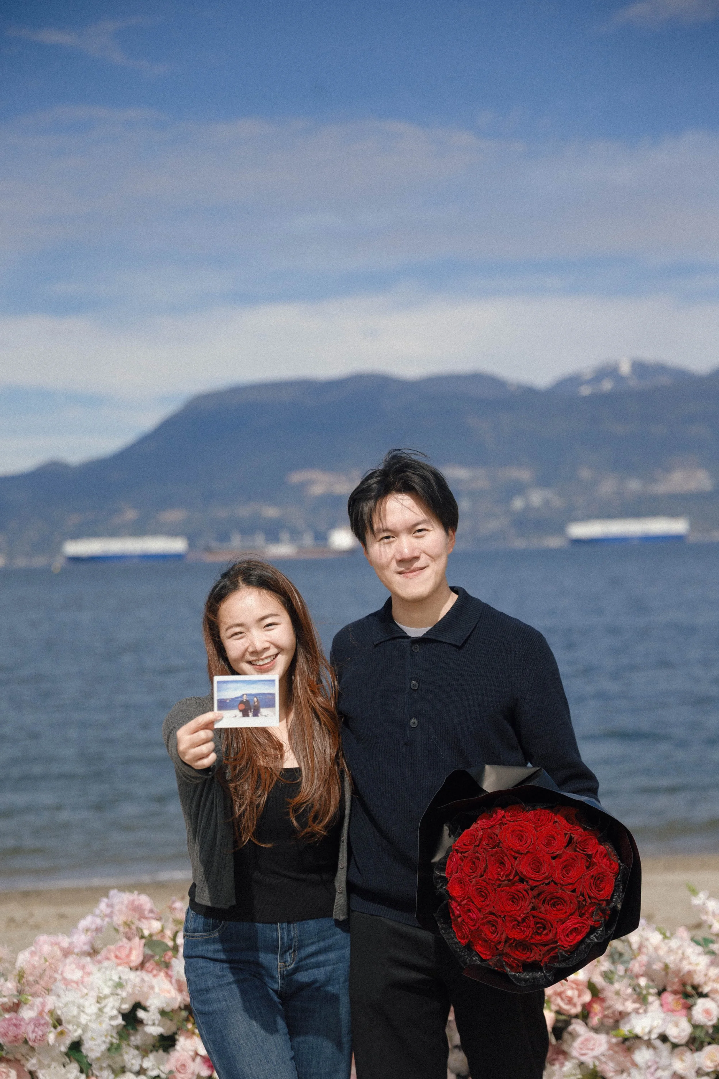 A woman smiling and holding a photo, standing next to a man who is holding a bouquet of red roses, at a location with a body of water and mountains in the background.