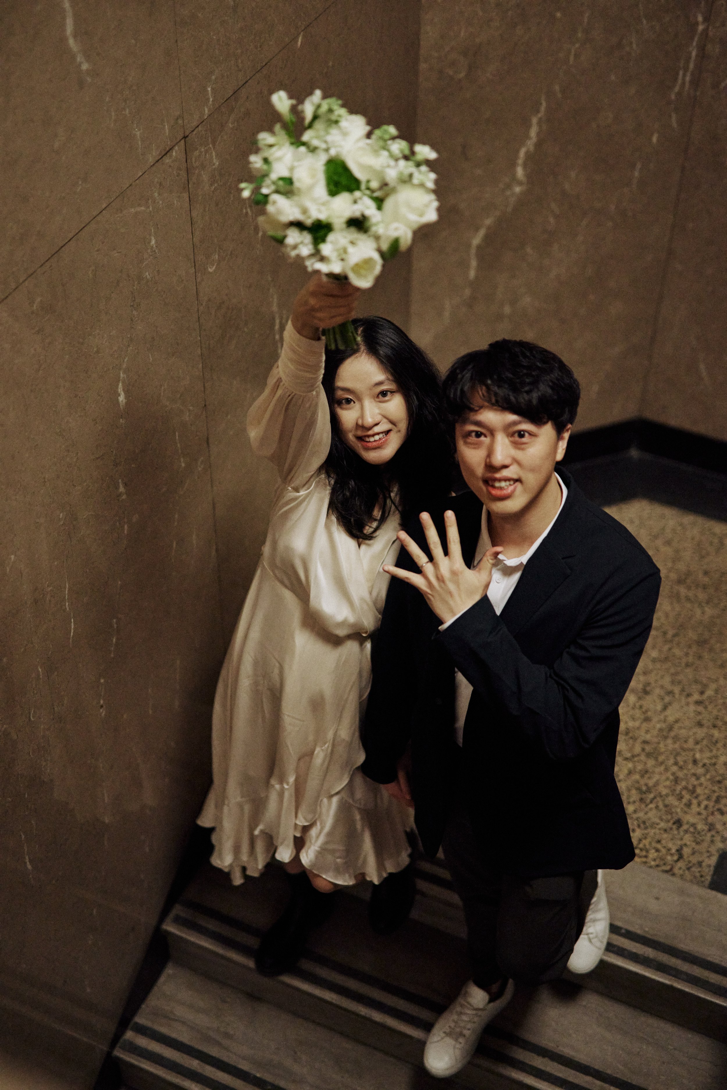 A woman with long dark hair wearing a cream-colored dress holding a bouquet of white flowers, standing next to a man with curly hair wearing a black blazer, white shirt, and white sneakers, both smiling and showing a hand with a ring.