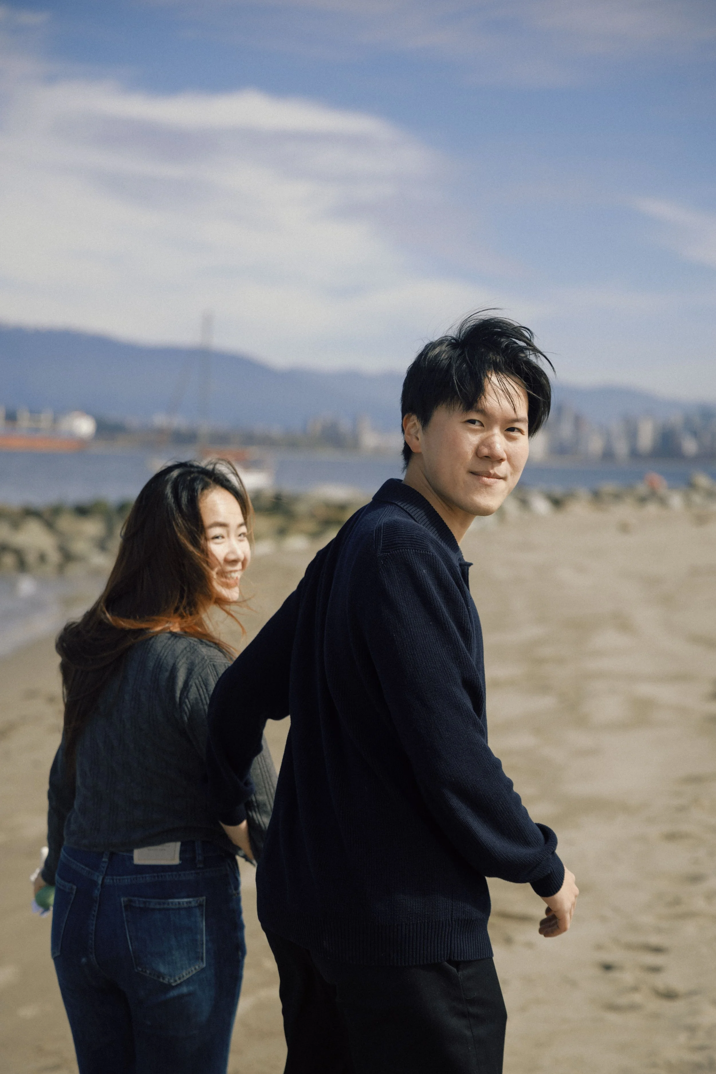Two young people, a man and a woman, are walking on a beach with a city skyline and mountains in the background. The man is looking at the camera with a slight smile, and the woman is smiling as she looks away from the camera.