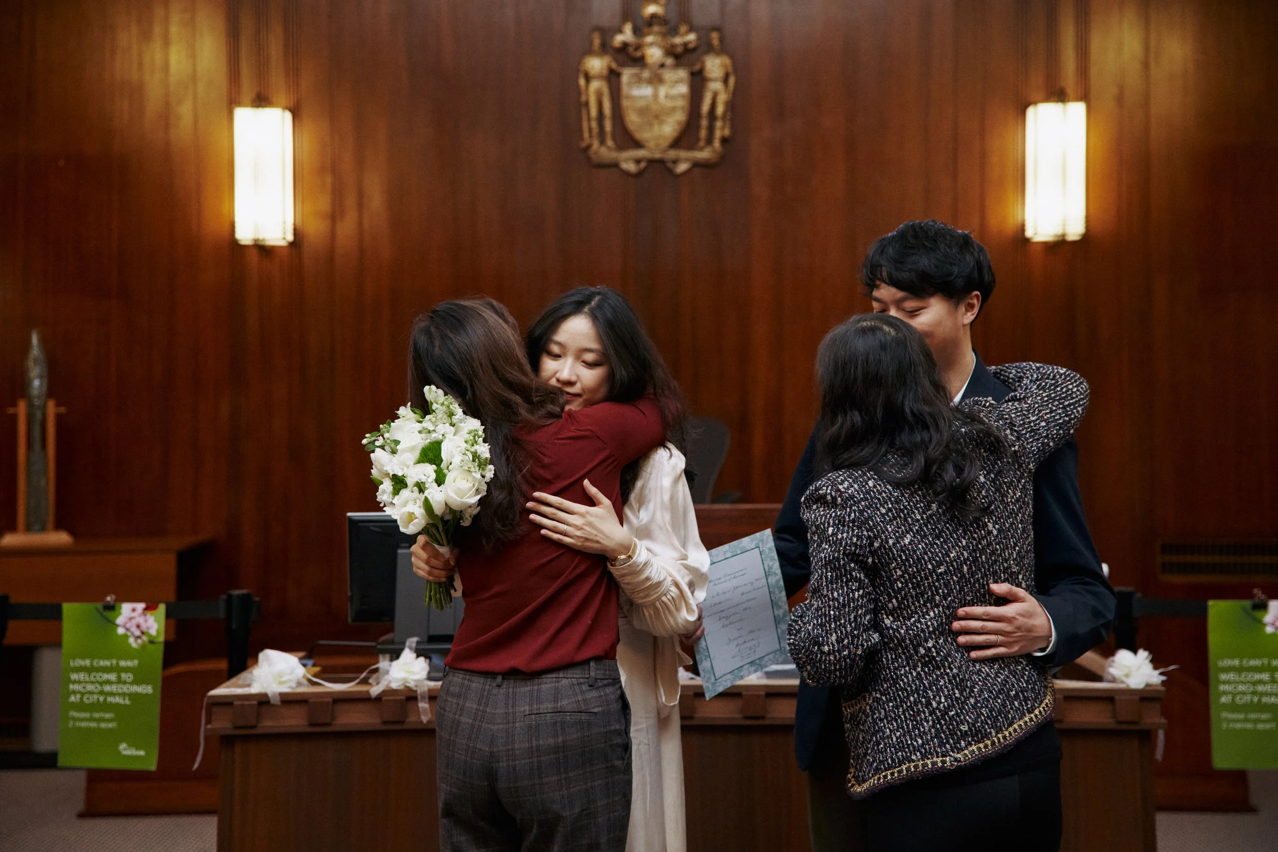 Four people hugging in a formal room with wooden walls and coat of arms above, two women and two men, one woman holding a bouquet of white flowers, signing documents on a table.