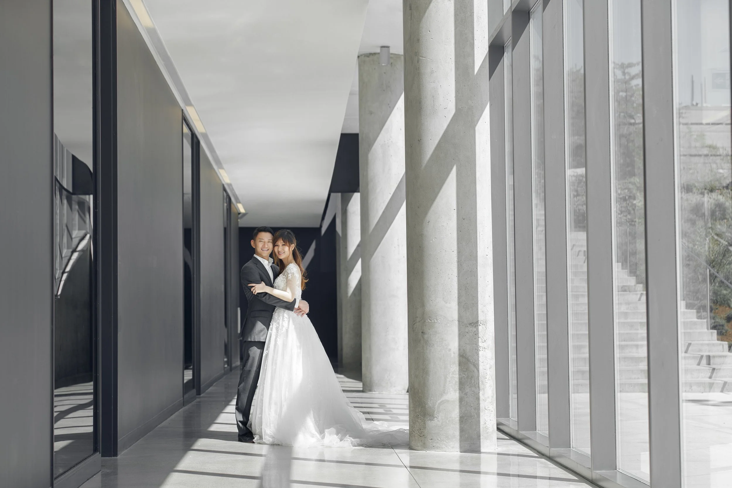 A smiling couple in wedding attire standing close together in a modern building with large floor-to-ceiling windows, concrete columns, and natural light.