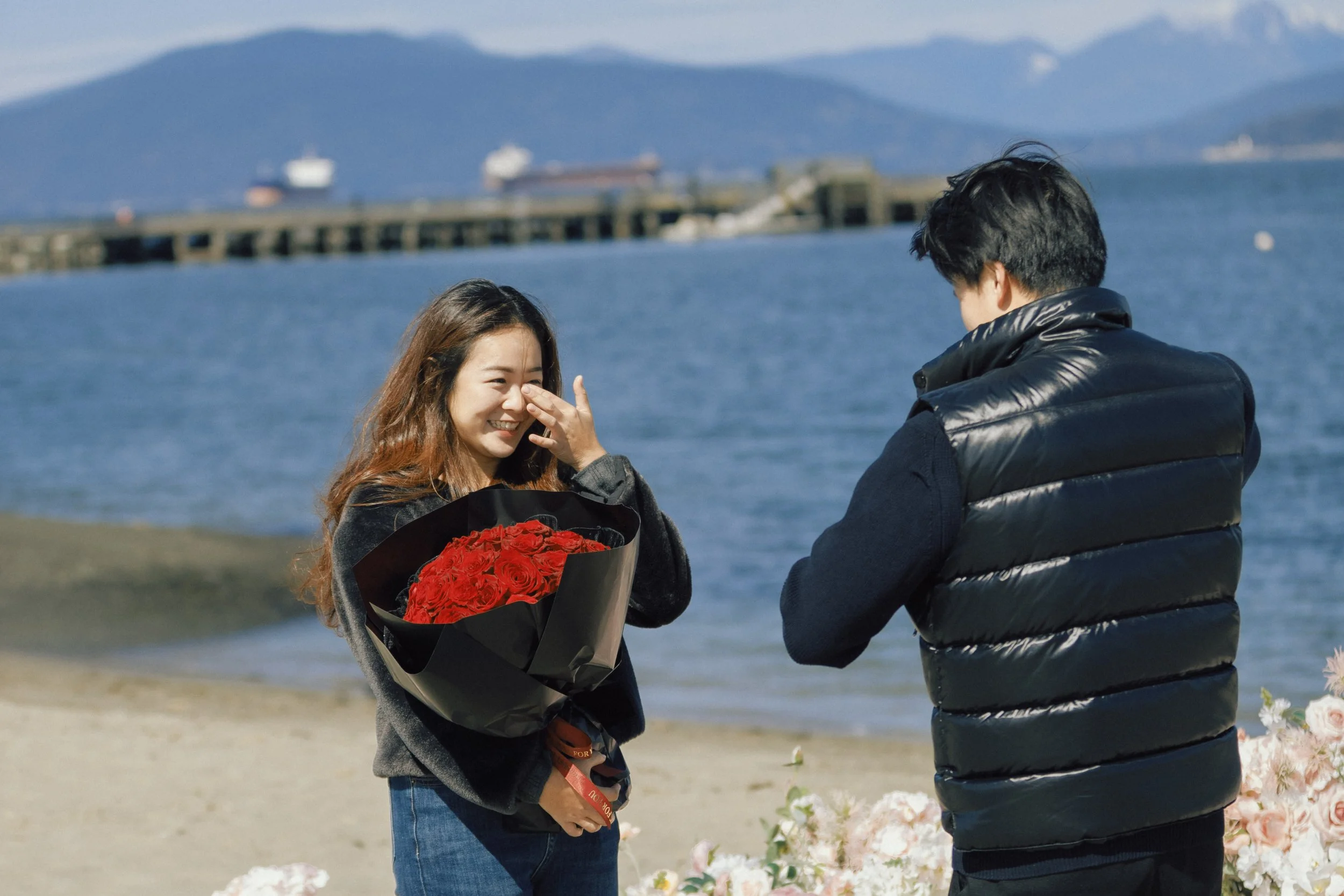 A man giving a woman a flower bouquet as she reacts happily, near the water with ships and mountains in the background.