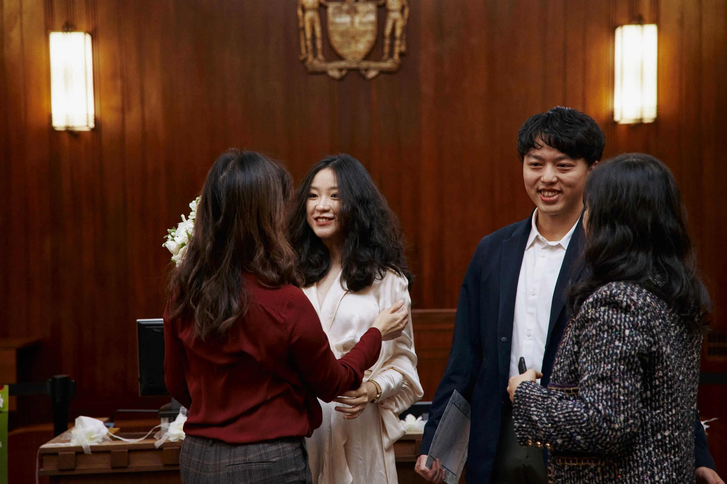 A group of four people in formal attire are gathered in a wood-paneled room with medals and an emblem on the wall, engaged in conversation and smiling.