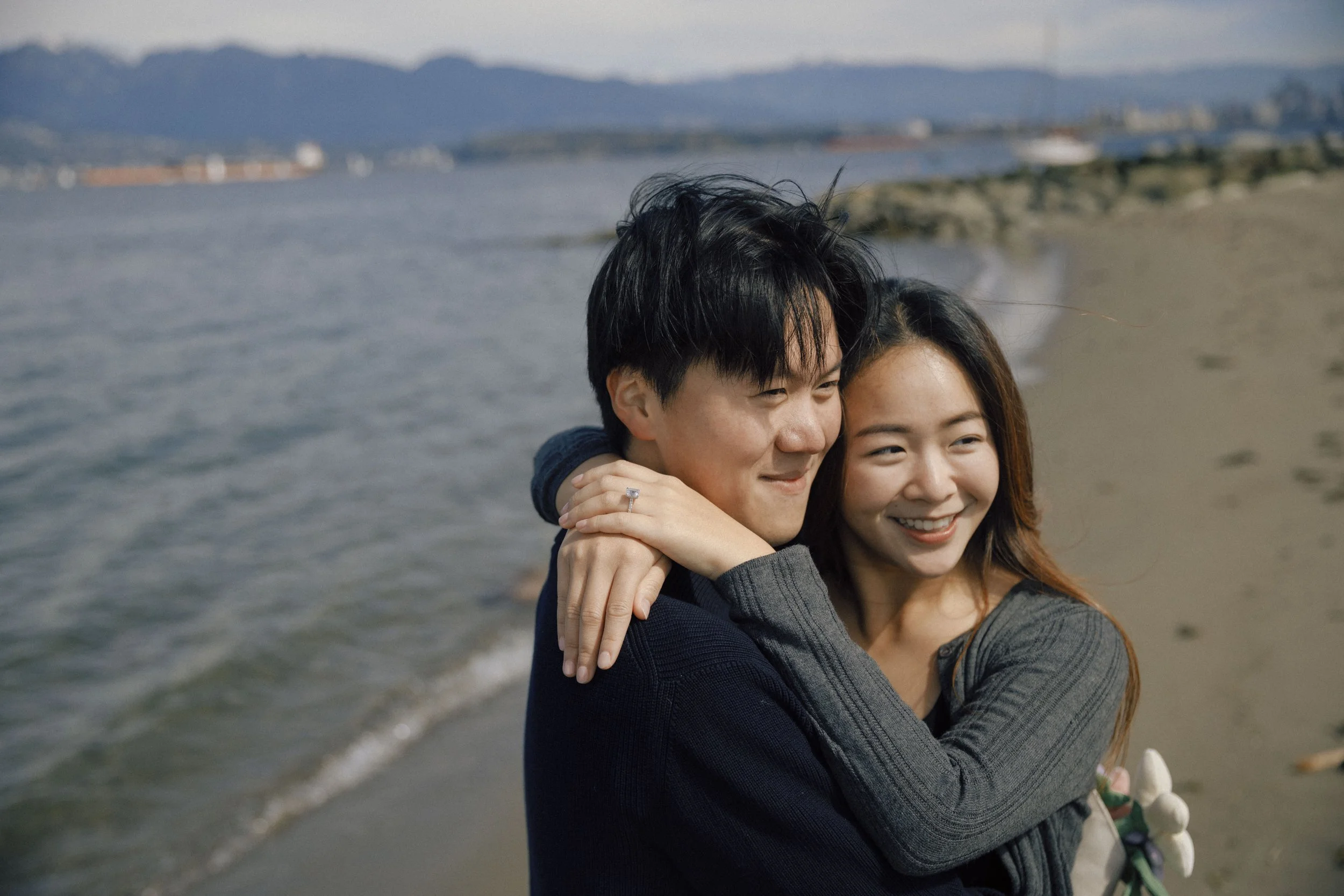 A young man and woman hugging on a beach, with mountains and water in the background.