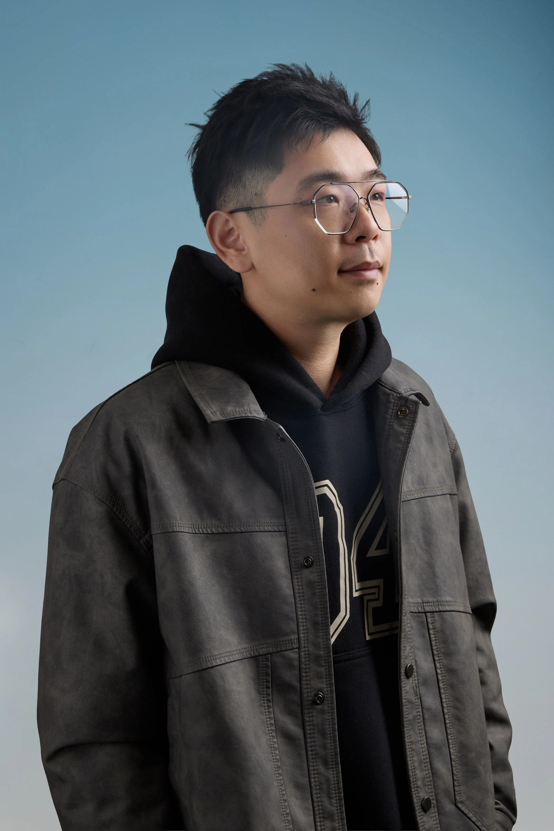 A young man with black hair wearing glasses, a black hoodie, and a gray jacket, looking to the side against a light blue background.