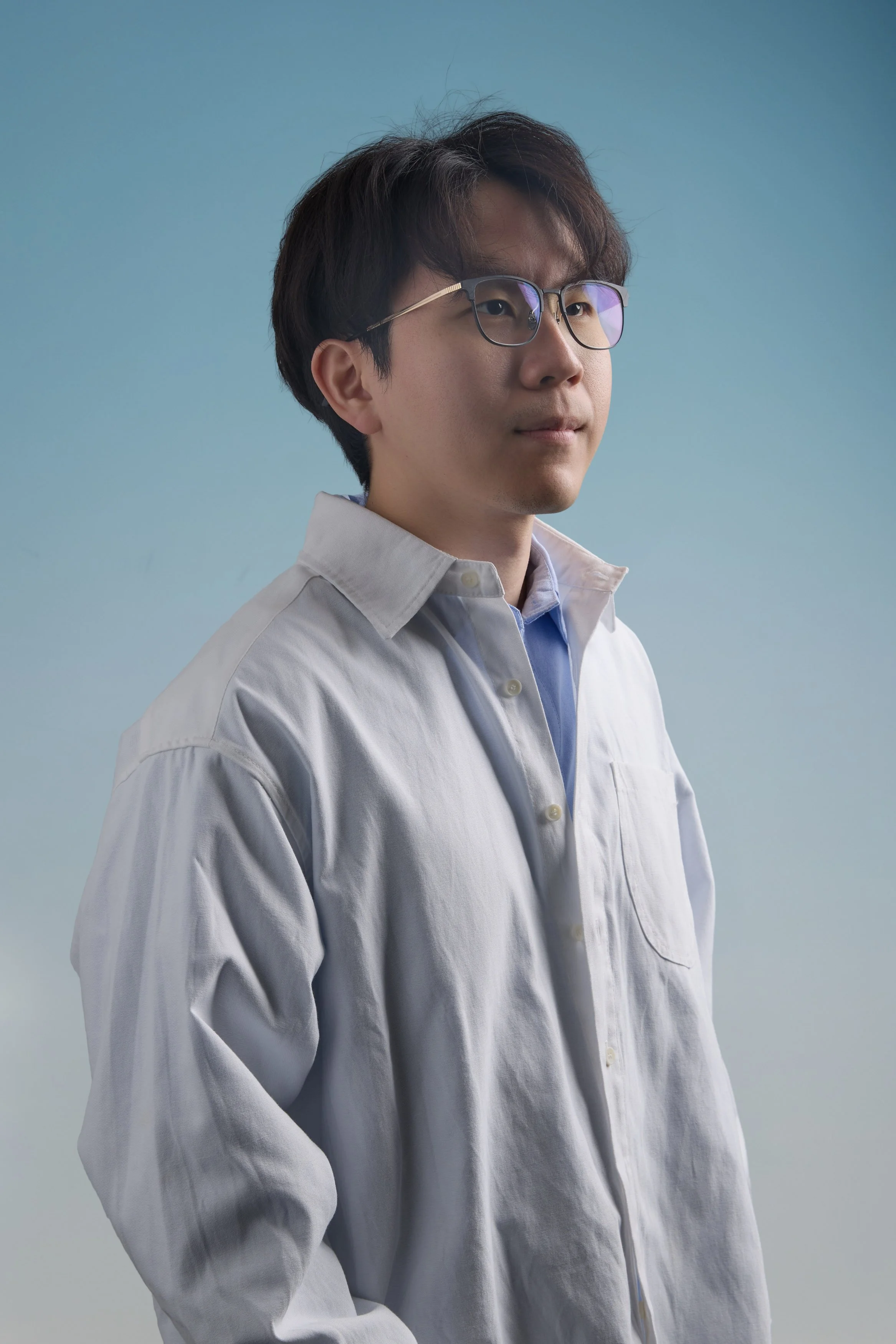 A young man with dark hair and glasses wearing a white shirt against a blue background.