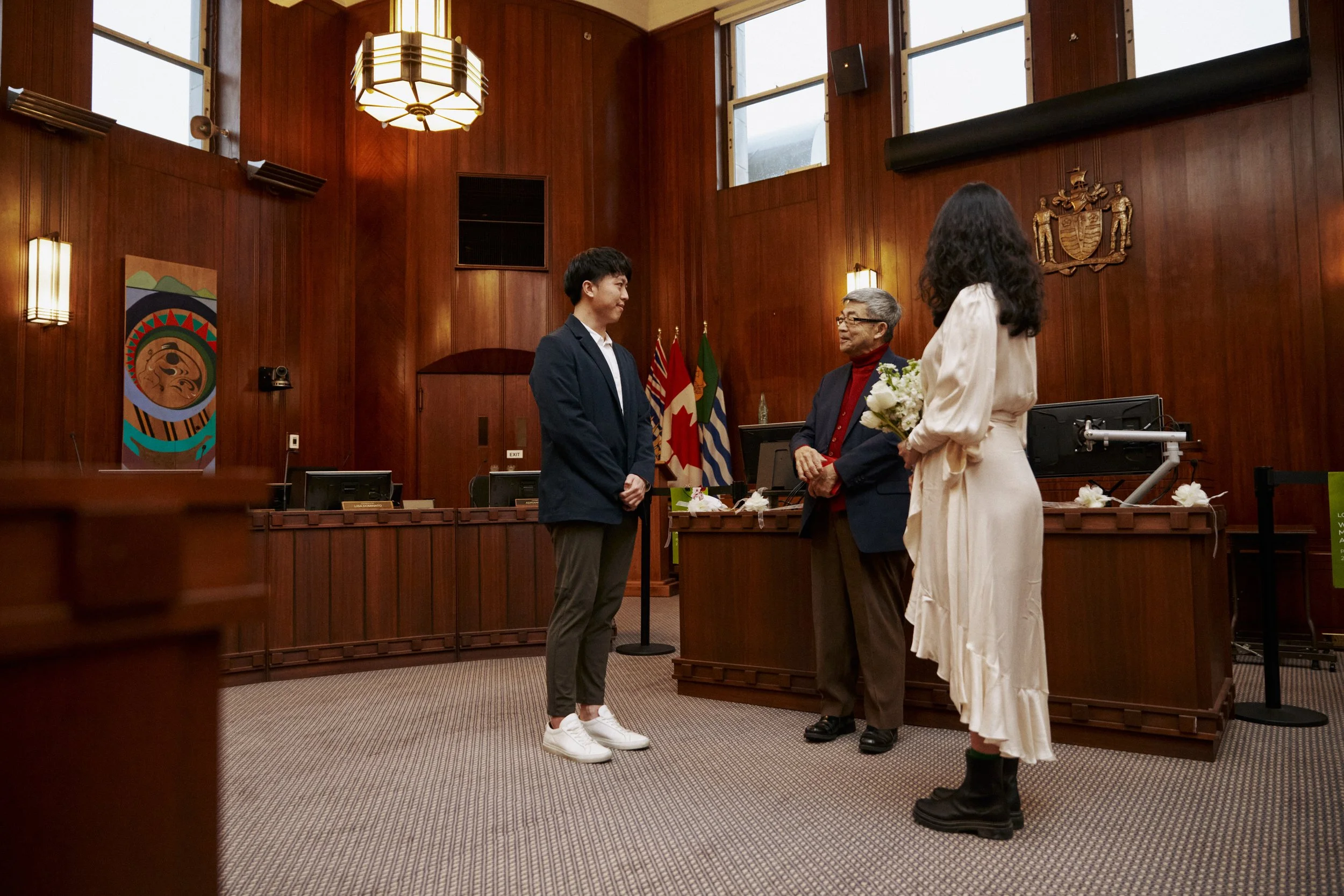A young man in a suit and sneakers stands in front of a judge and a woman dressed in a white gown in a wood-paneled courtroom with flags and crest behind them, engaged in a ceremony.