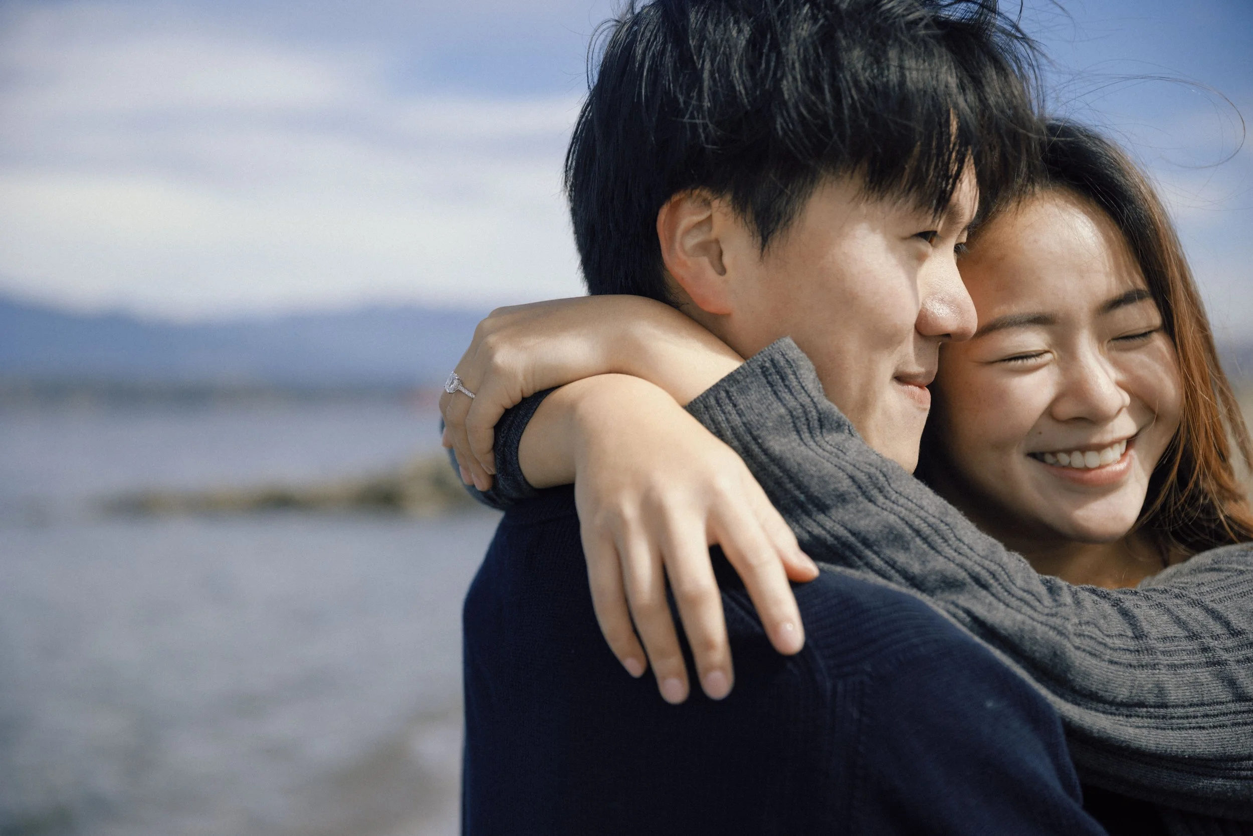 Two people hugging outdoors by the water, one with dark hair and the other with light brown hair, smiling with eyes closed, happy.