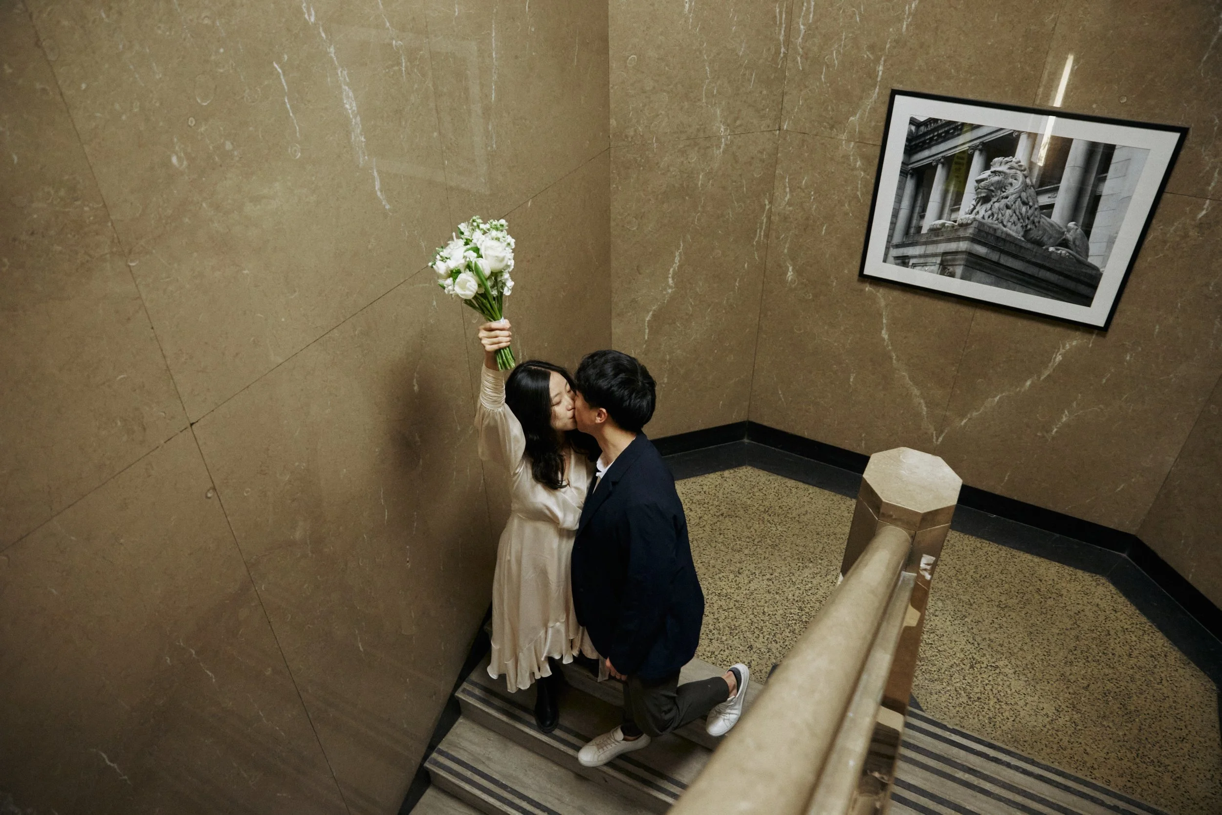 A couple sharing a kiss on a staircase in an indoor setting. The woman is holding a bouquet of white flowers and is dressed in a light-colored dress. The man is wearing a dark jacket and pants. There is a framed black-and-white photograph of a lion s