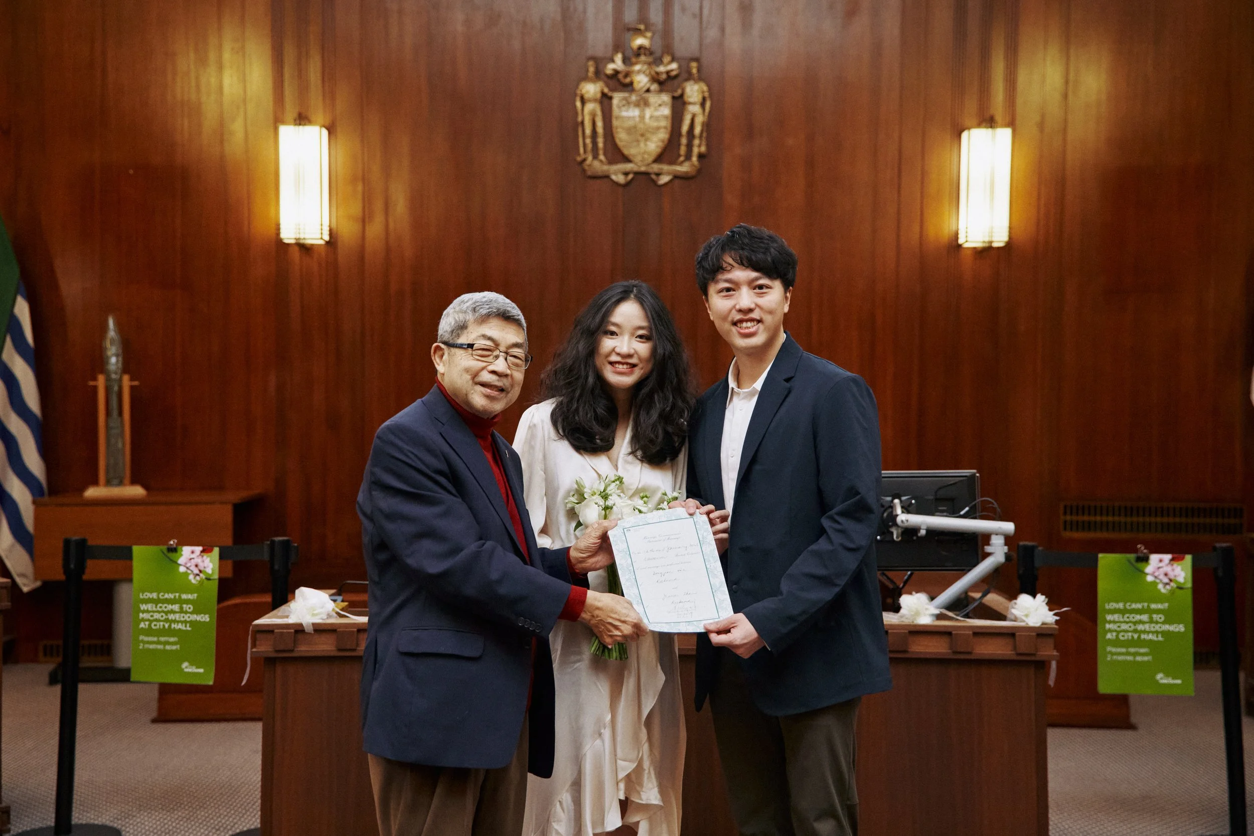 Three people standing together in a formal setting, with a wooden wall and crest behind them. An older man in glasses wearing a dark suit and red shirt is holding a bouquet of white flowers and a certificate, with a young woman in a white dress and a