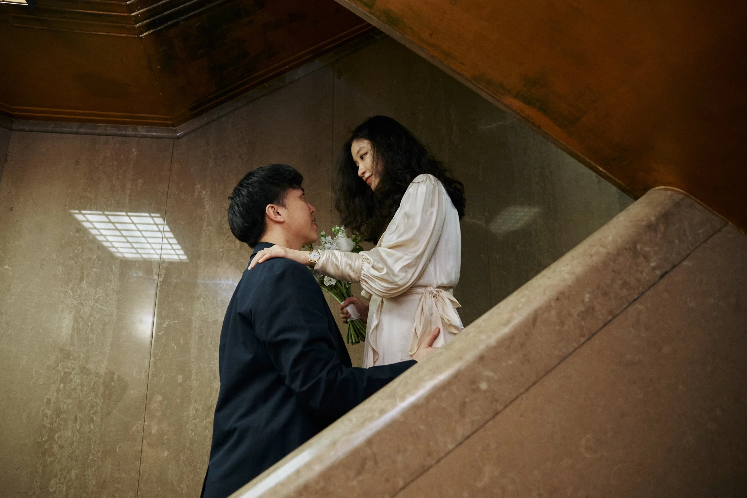 A man kneels on one knee holding a bouquet of flowers while a woman stands above him, smiling and looking into his eyes. They are on a staircase with a marble wall background, and the woman is dressed in a light-colored satin robe.