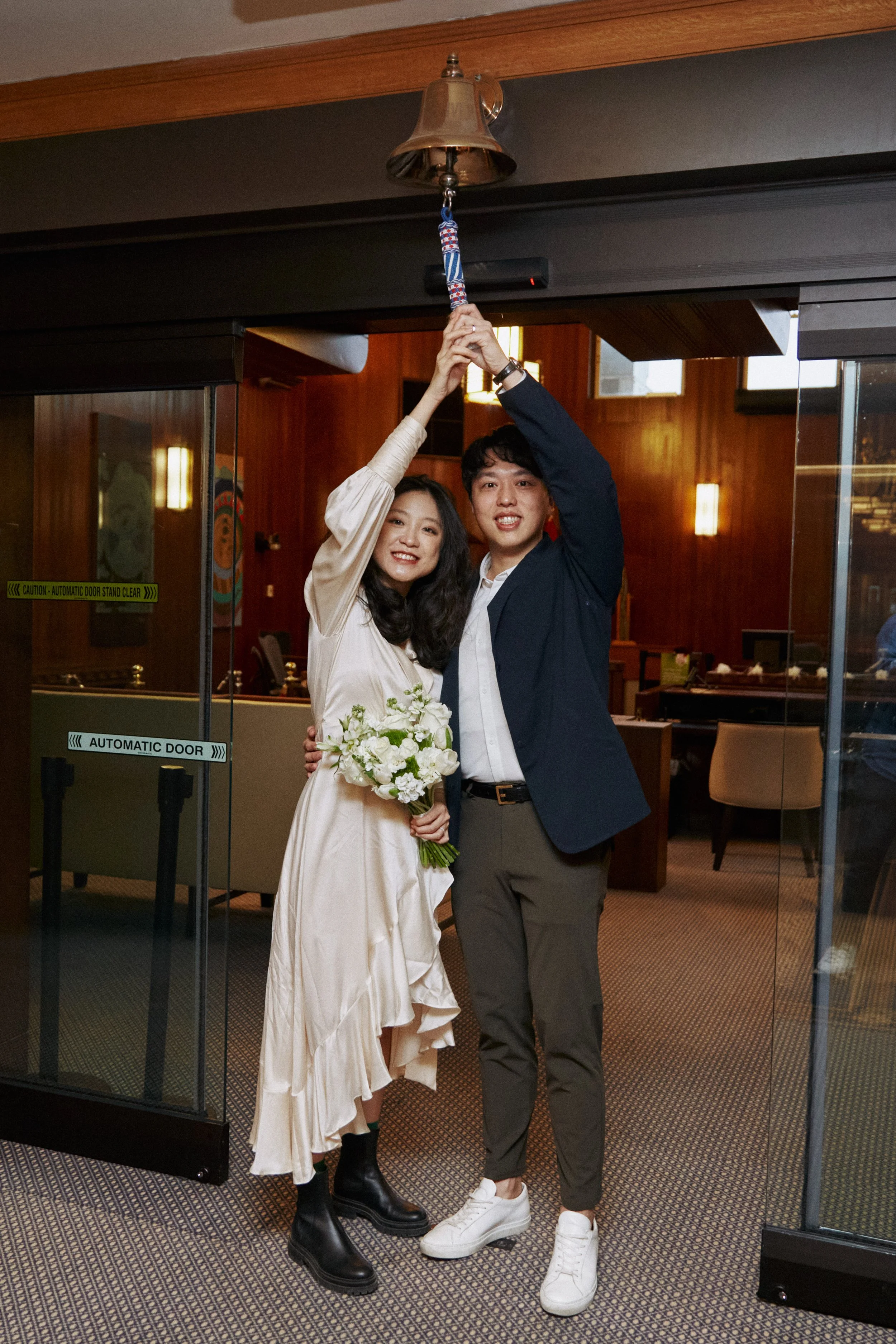 A young couple is celebrating in a restaurant. The woman is holding a bouquet of white flowers and is smiling. The man is smiling, wearing a navy blazer, gray pants, and white sneakers. They are both holding a bell above their heads.