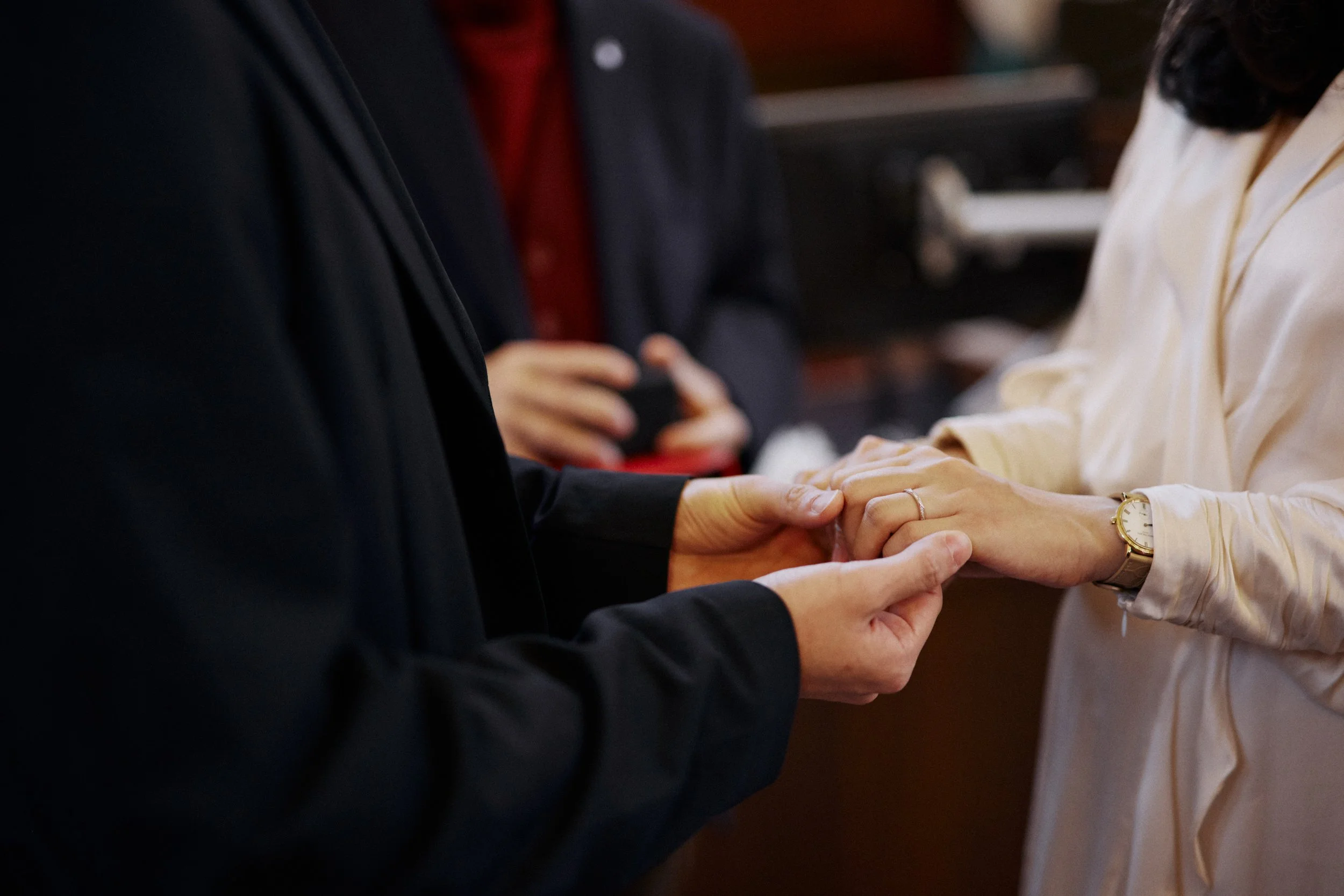 Close-up of two people holding hands during a wedding ceremony, with a third person in the background using a phone.