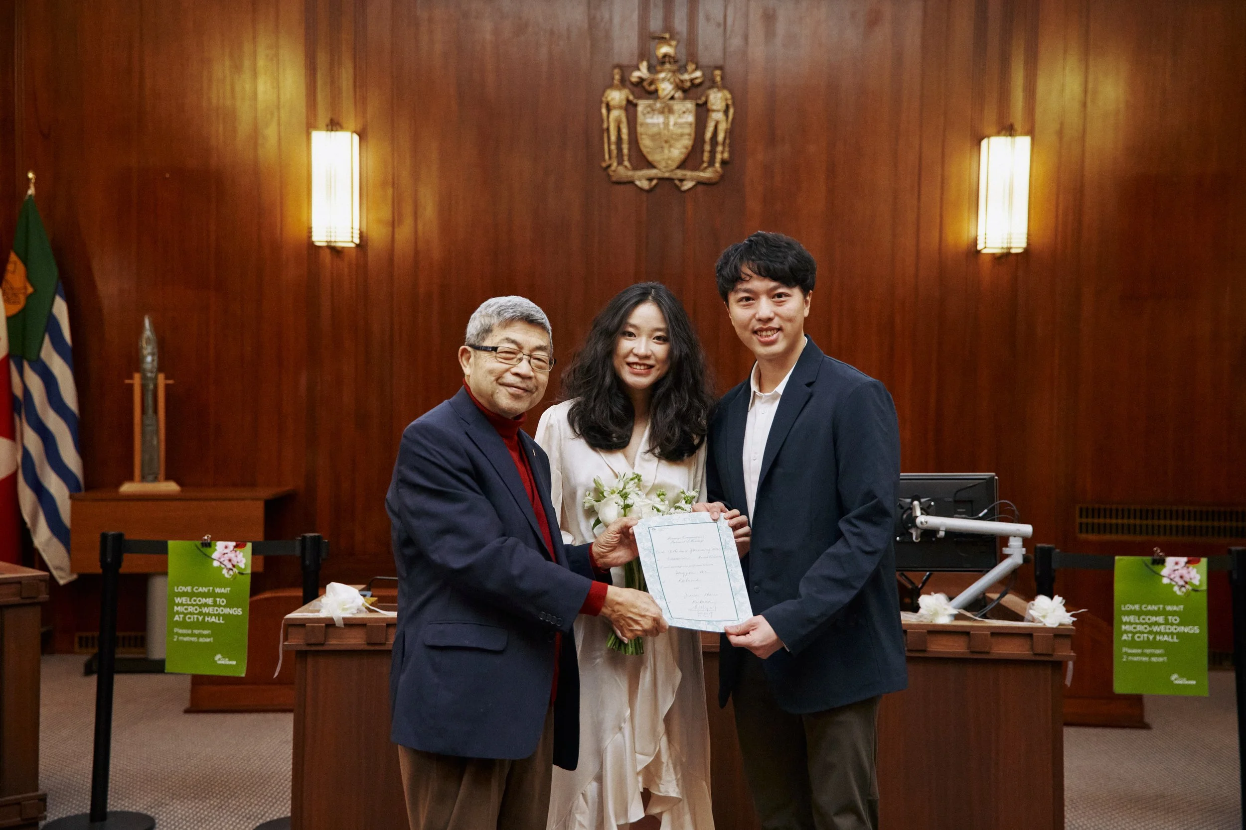 A woman receiving an award from an older man with glasses in a formal setting, with a young man standing beside her. Wooden walls, flags, and signs are visible in the background.