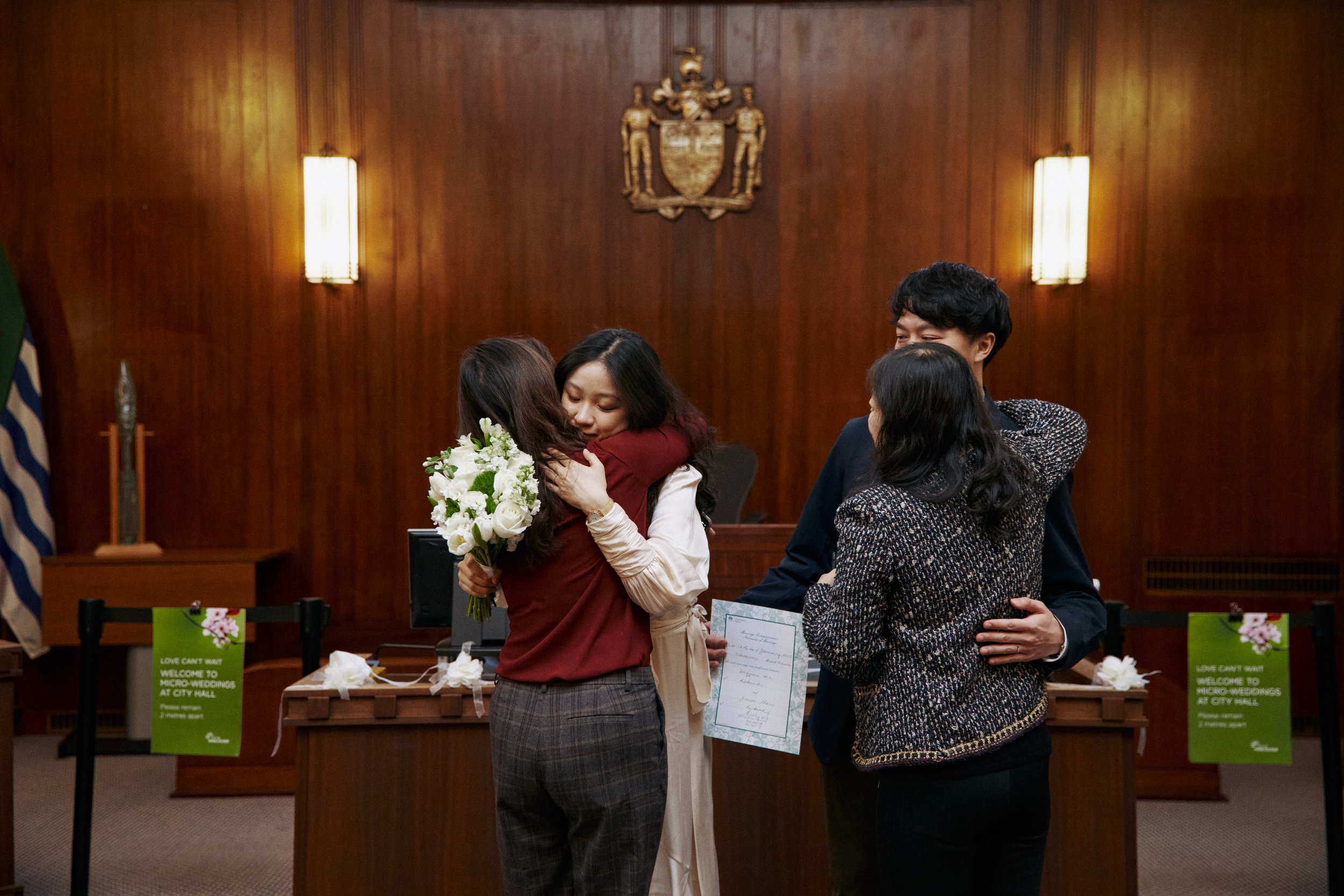Group of four people hugging in a formal room, with a wooden wall background and two wall-mounted lamps.