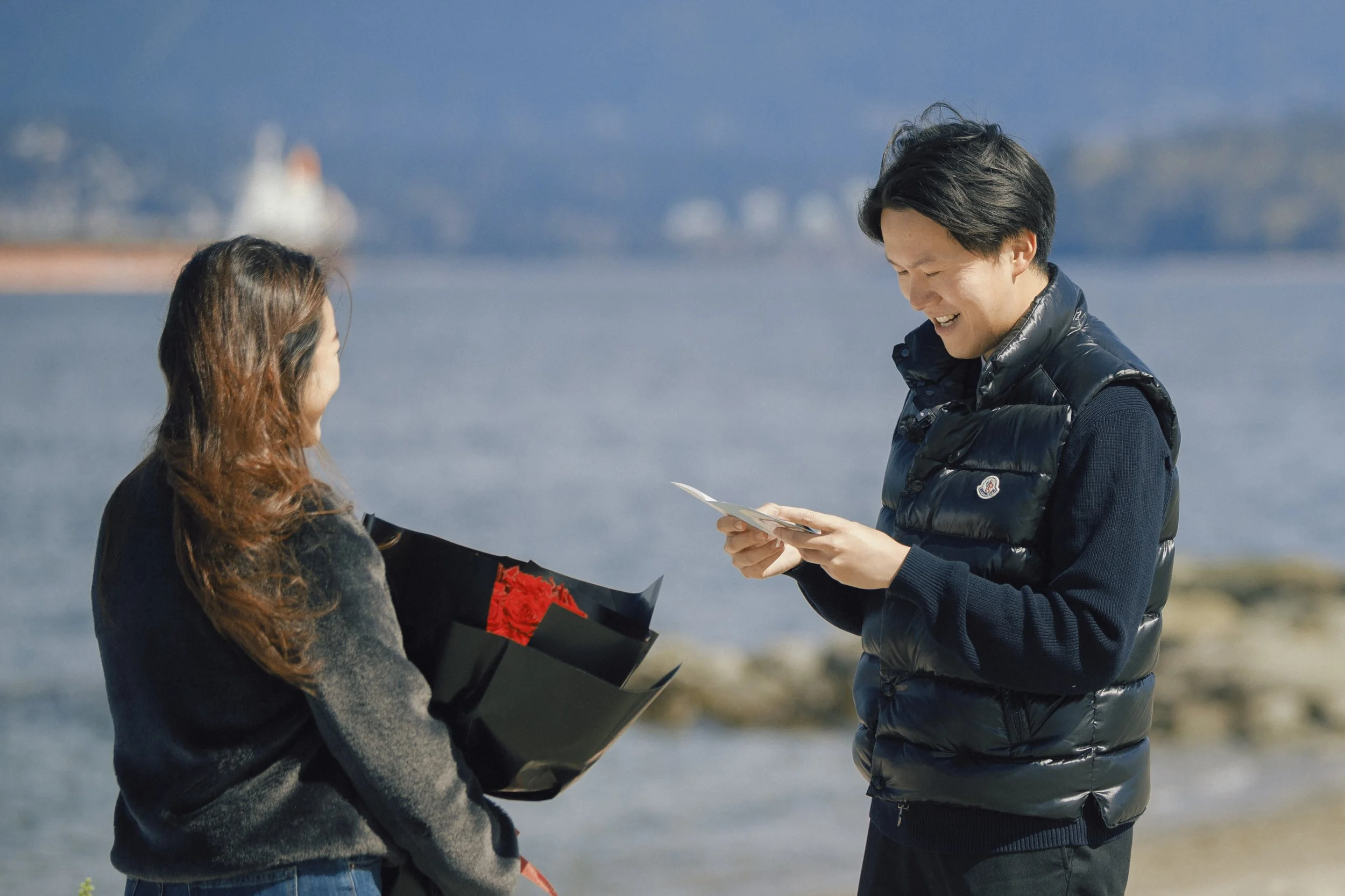 A man giving a woman a certificate or paper, with a woman holding a bouquet of red roses, standing near water with a blurred ship in the background.
