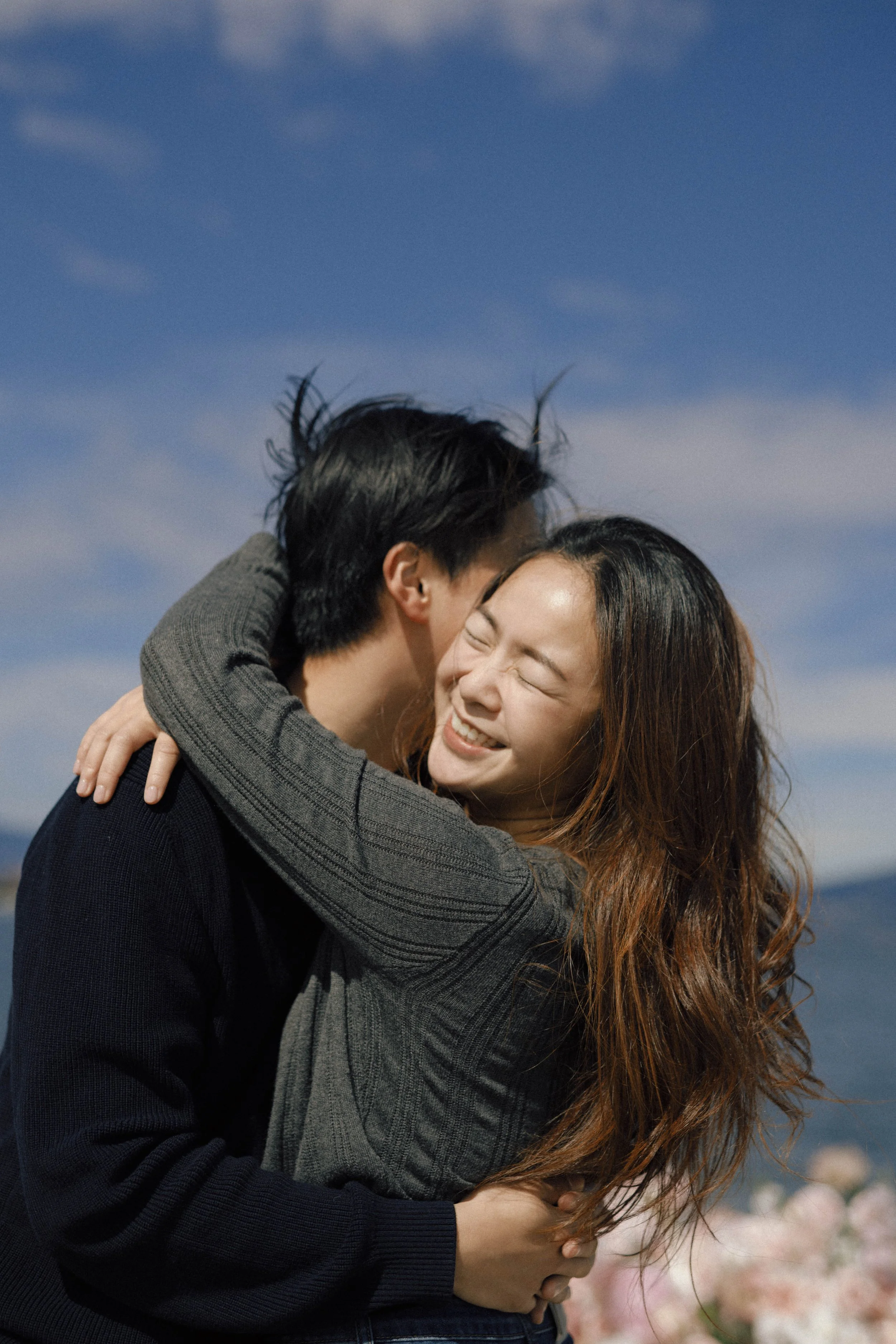A couple embracing outdoors, smiling with eyes closed against a blue sky, with pink flowering trees in the background.