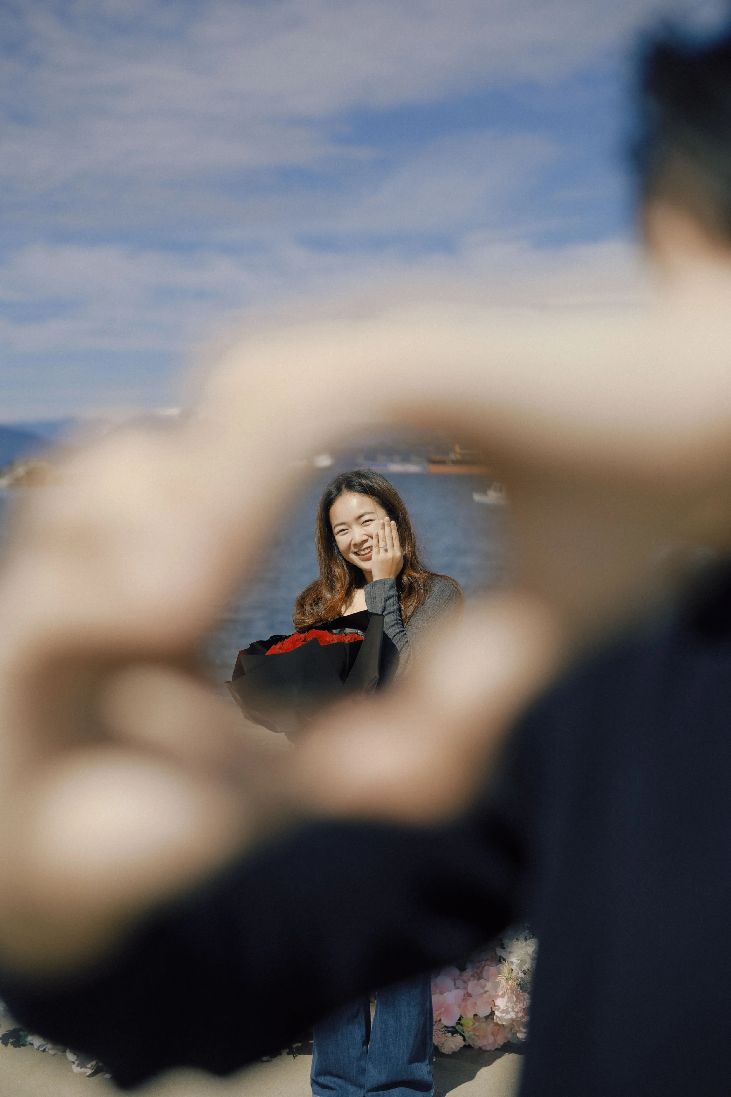 A smiling woman with long brown hair, holding a bouquet of red flowers and covering part of her face with her hand, with a waterfront and boats in the background, viewed through blurred foreground objects.