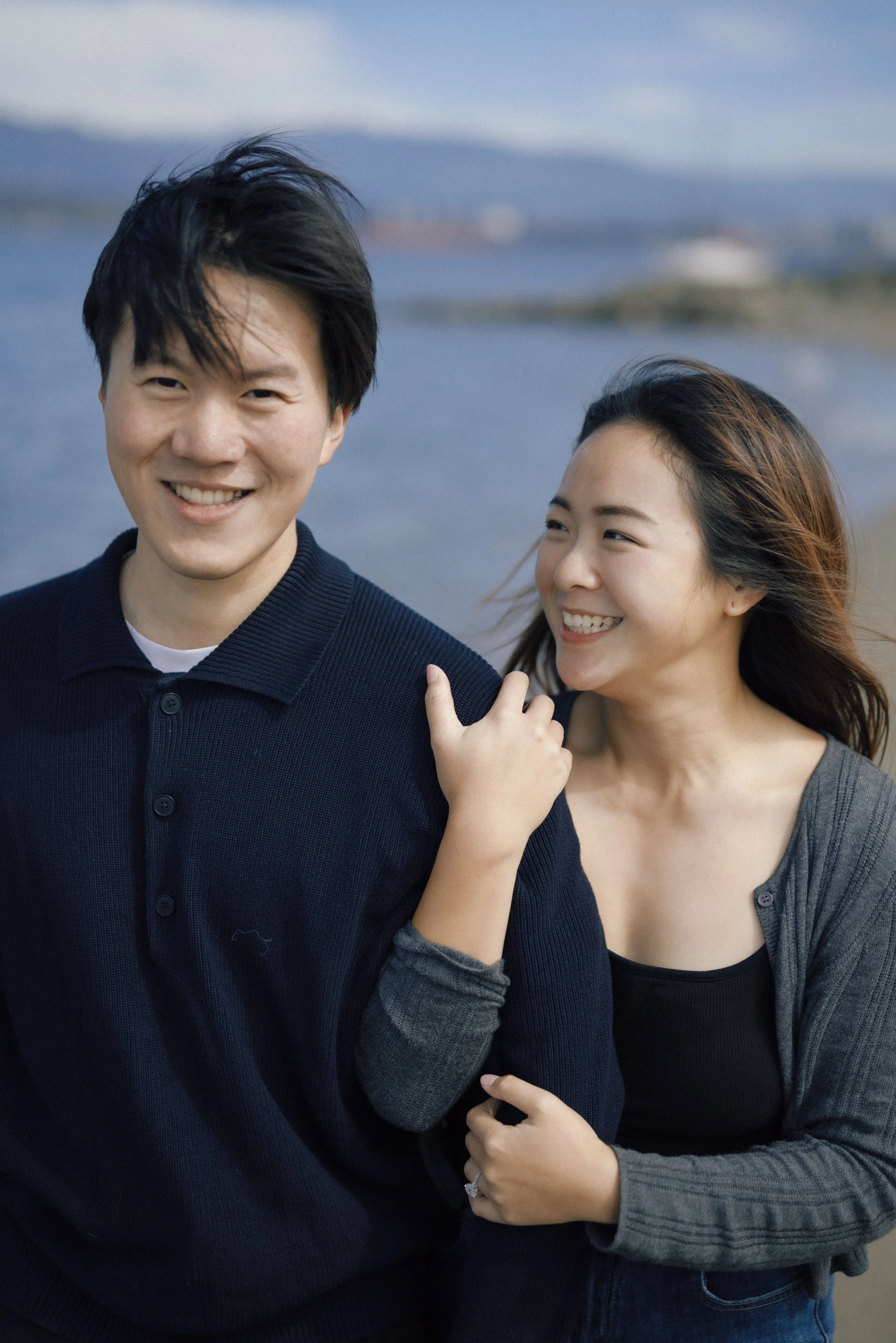 A smiling Asian man and woman at the beach. The woman's hand rests on the man's arm, and they are both dressed casually, with a blurry shoreline and mountains in the background.