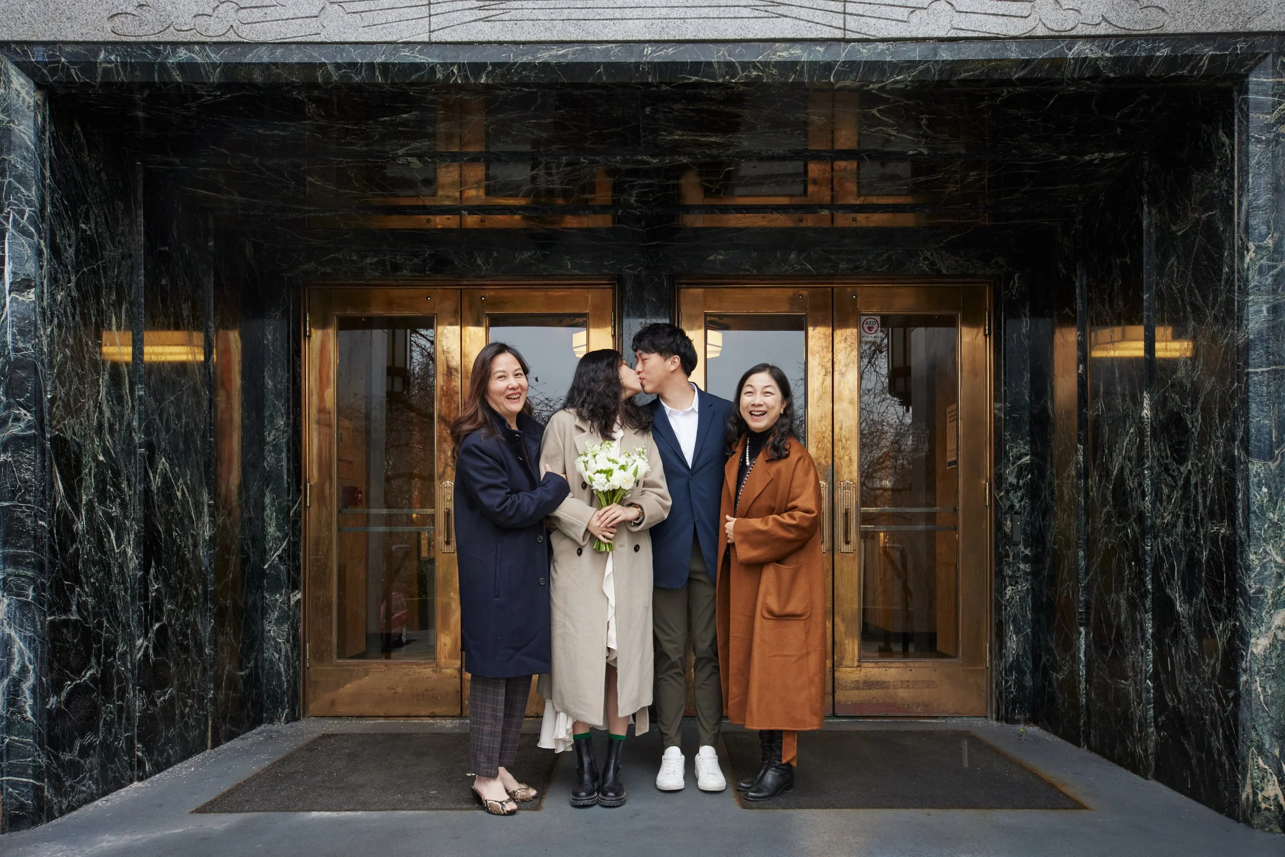 Four people standing in front of a building entrance, with a woman holding a bouquet of white flowers and the others smiling and kissing. The building has black marble walls and glass doors with brass accents.