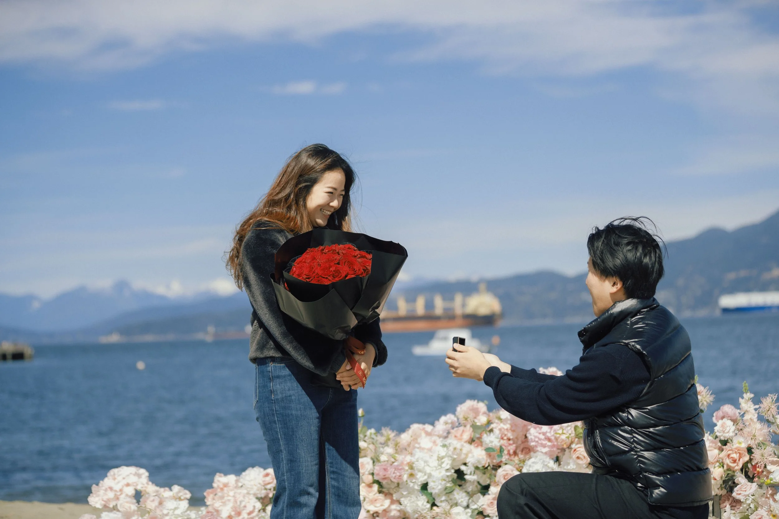 A woman is receiving a bouquet of red roses from a man by the water with ships and mountains in the background, surrounded by pink and white flowers.