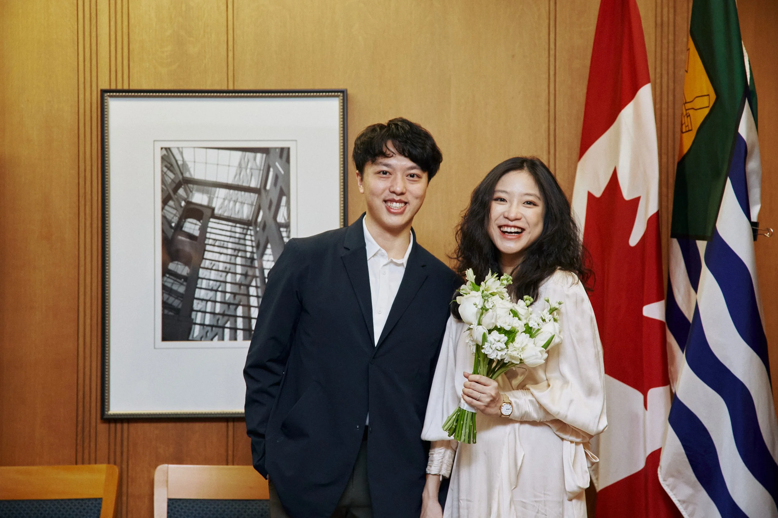 A young man and woman standing together indoors, smiling. The woman is holding a bouquet of white flowers. Behind them are several flags, including the Canadian flag and the Greek flag, and a framed black-and-white photograph of architectural structu