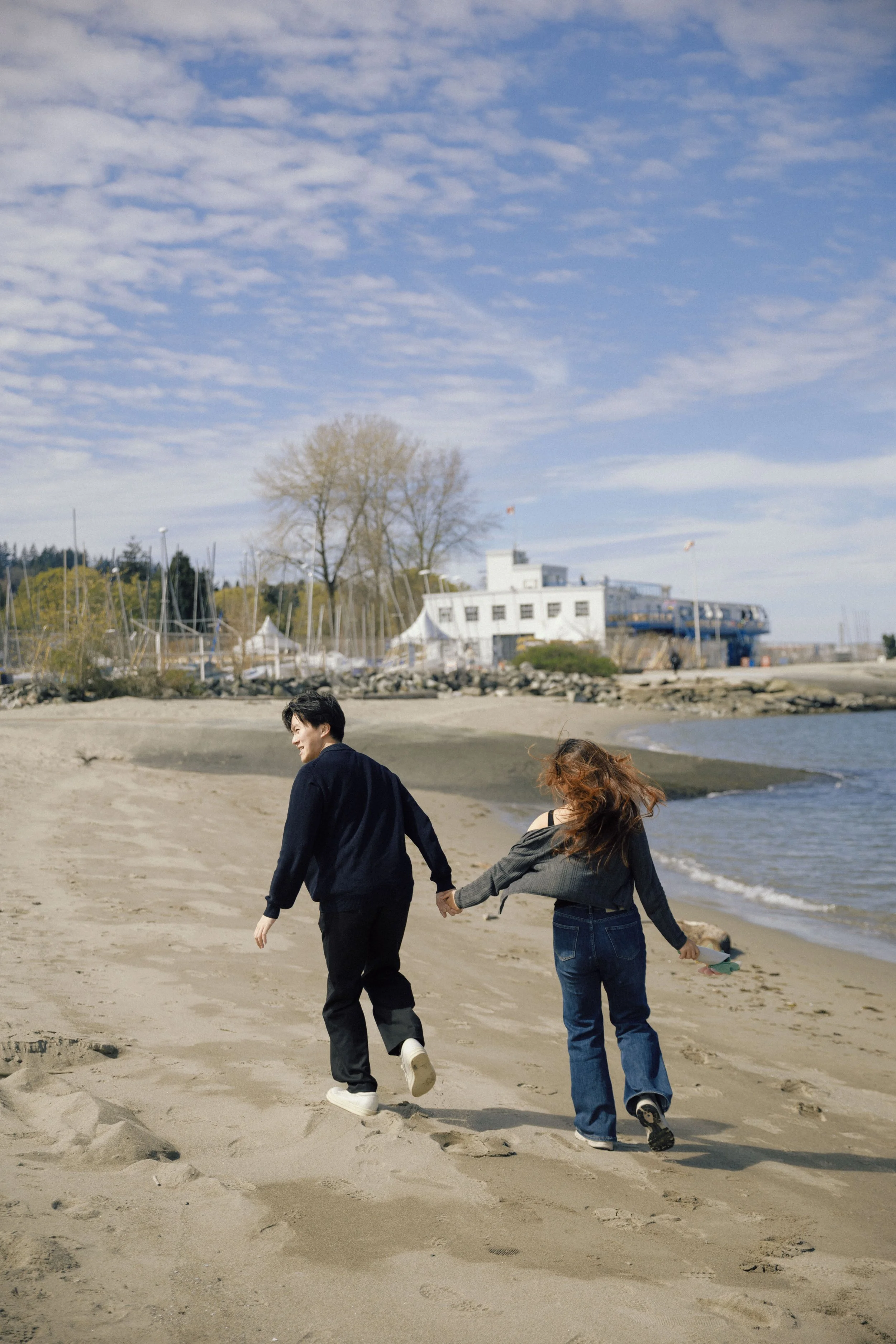 Two people holding hands and walking on a sandy beach with sailboats and a white building in the background under a partly cloudy blue sky.