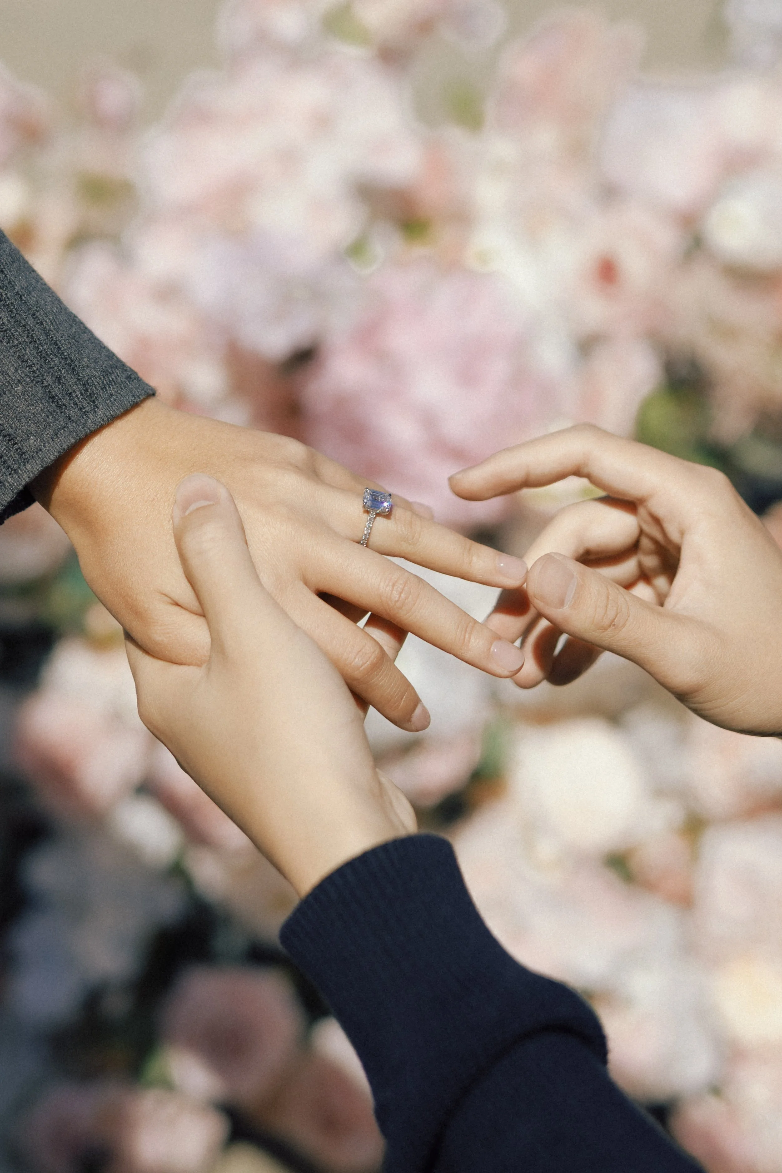 A person is putting a ring on another person's finger during a wedding ceremony or engagement moment, with a blurred background of pink flowers.