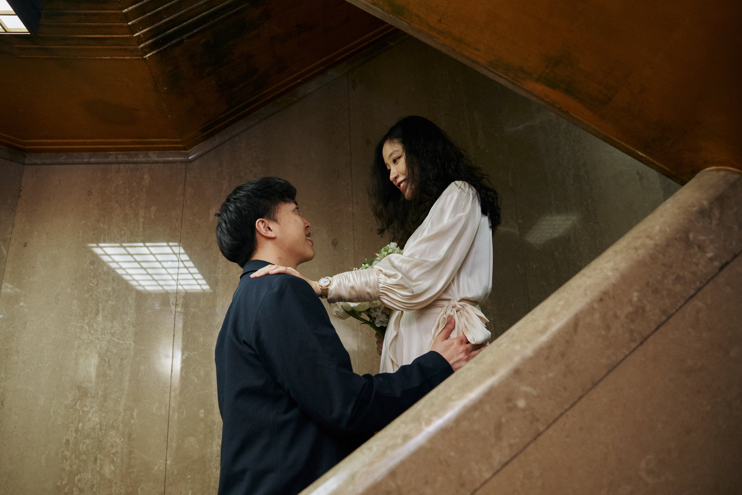 A man and woman sharing a moment on a staircase. The man, dressed in a dark suit, is kneeling and looking up at the woman. The woman, wearing a light-colored dress, is smiling and holding a bouquet of flowers. They are in an indoor setting with polis