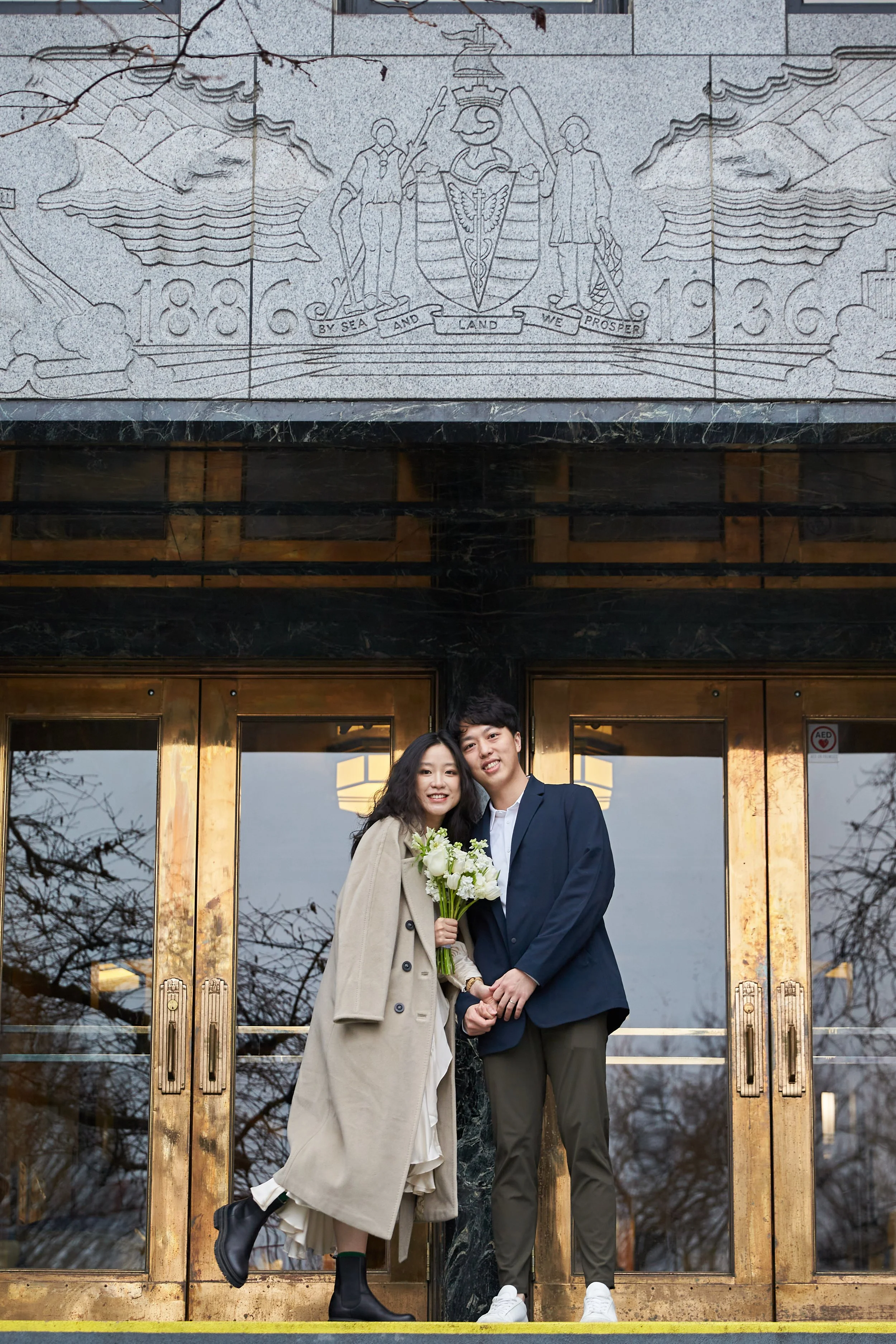 A happy couple stands in front of a large, glass door with gold-colored framing. They are smiling and holding hands, with the woman holding a bouquet of white flowers. The man is dressed in a dark suit, and the woman is wearing a beige coat and boots