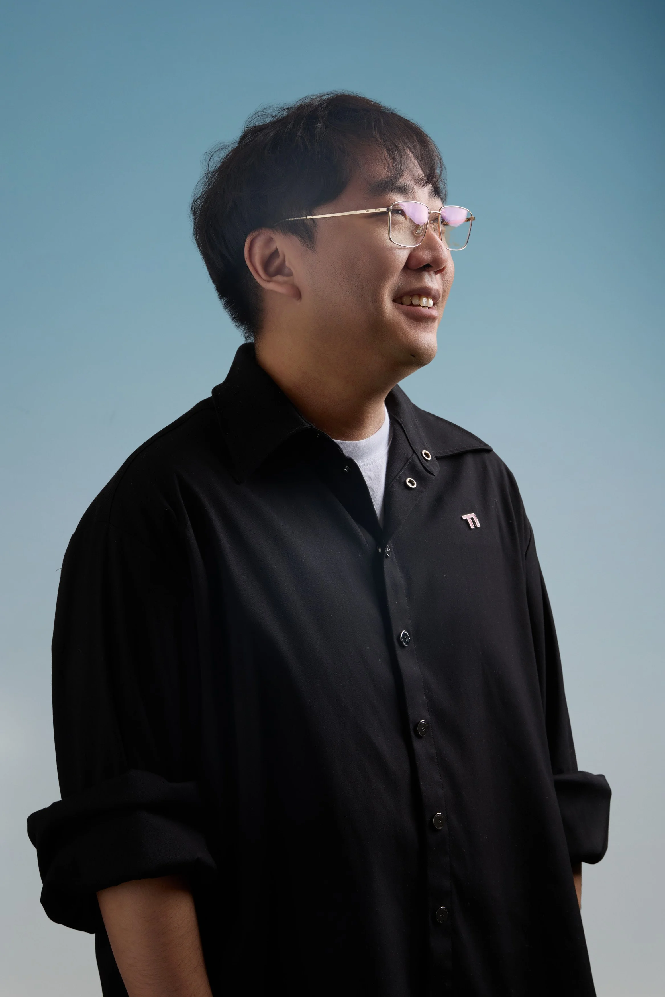 A middle-aged man with black hair, glasses, and a smiling expression, wearing a black button-up shirt, standing against a plain blue background.
